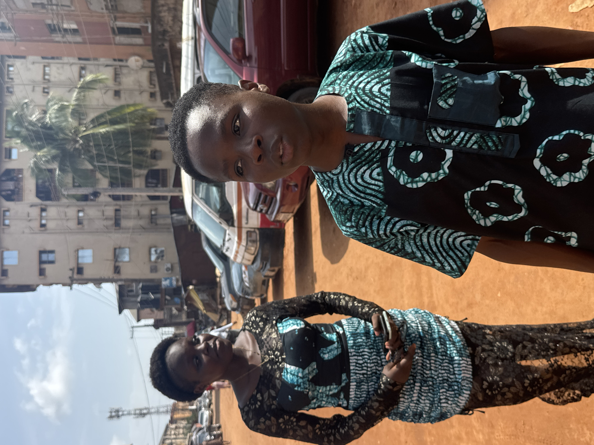 Two people are positioned in the foreground of an outdoor scene in Onitsha, Nigeria. The person in the top-center wears a black patterned top with teal and white markings and a black-teal collar. The second person, positioned below and to the left, wears a dress with black lace sleeves and a bodice patterned in teal, white, and black. They hold a rectangular object. Various vehicles are parked in the background, including a burgundy car partially visible behind the person in the top-center, a white and orange van, and other cars further back. Buildings are visible in the background, including a multi-story building with a palm tree situated in front of it. The ground appears to be reddish-brown. The sky is visible in the upper left corner and is pale blue.