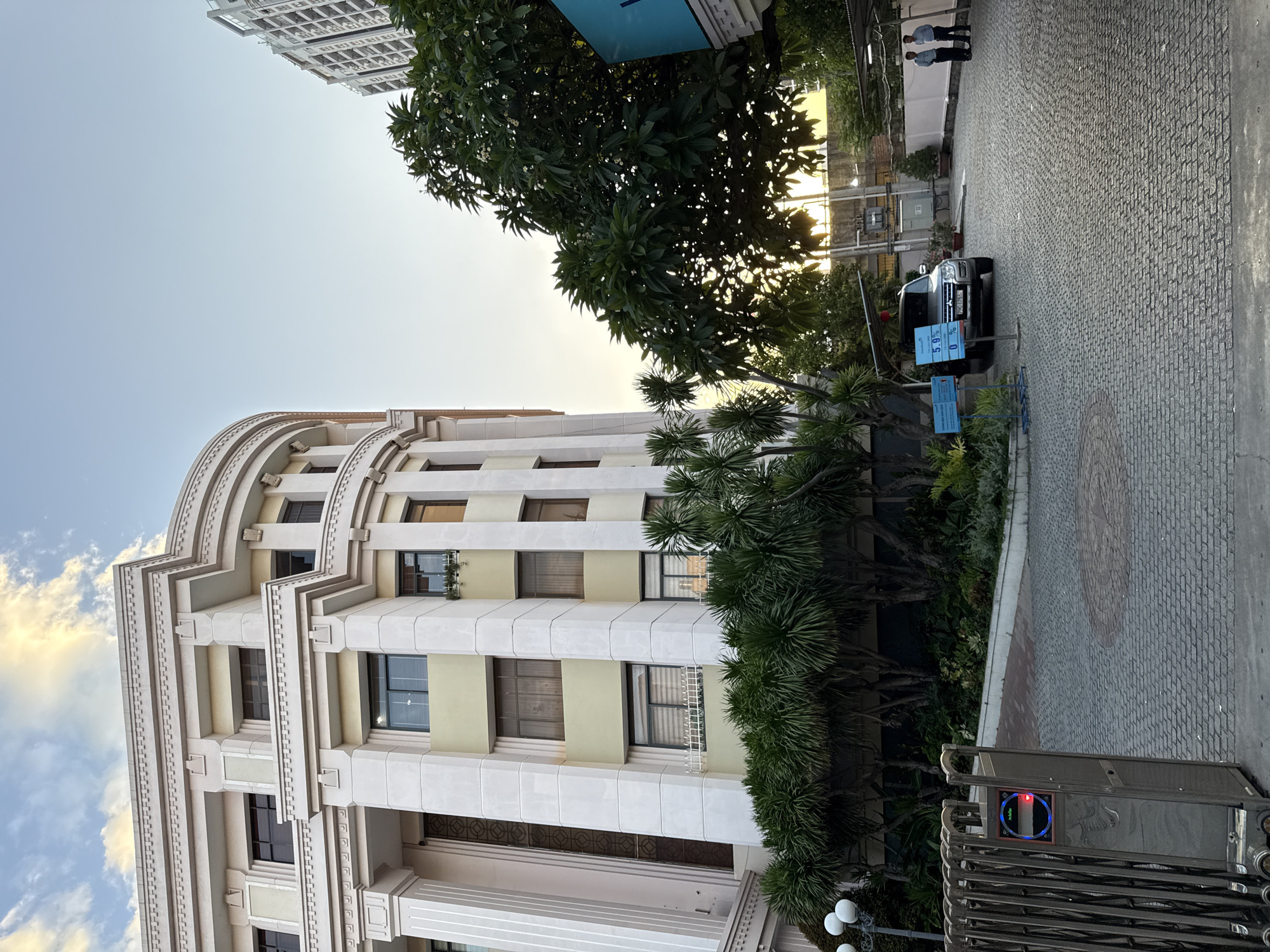 The image shows a street scene in Nha Trang, Vietnam. The foreground features a gray cobblestone driveway and a metal gate with an access control panel. To the left is the facade of a modern, light-colored building with multiple floors, arched windows, and decorative elements. In the middle ground, a dark-colored SUV is parked on the driveway, and a blue sign with text and a percentage display is visible near it. Several trees and lush green foliage are present throughout the scene. In the background, two men in casual attire stand near a white wall, and a tall, modern building is visible on the right side of the frame. The sky is a clear blue with some scattered clouds, and the lighting suggests it is late afternoon or early evening.