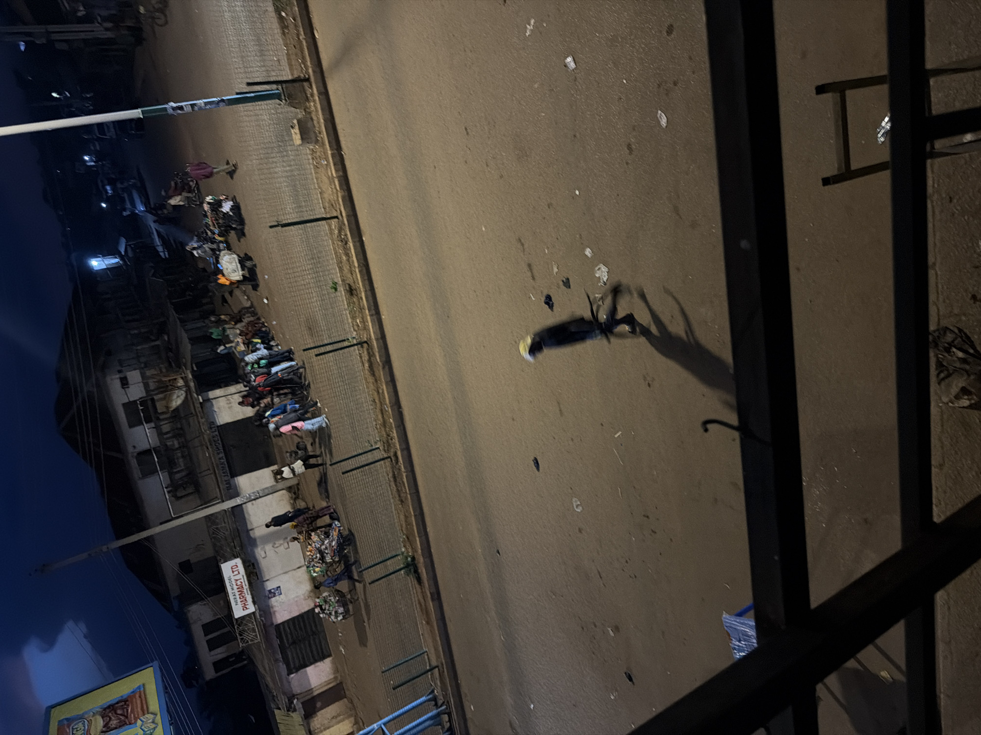 An elevated view captures a street scene at night in Bukuru, Nigeria. The street is paved with cobblestones and asphalt, with scattered debris and litter visible on the road surface. Several individuals are present, some walking, others gathered in small groups.
In the foreground, a lone figure wearing a dark garment and a yellow cap walks across the street, casting a long shadow. The figure appears to be moving from left to right.
Along the side of the street, a row of buildings lines the left side of the image. Some of these buildings appear to be shops, with signs visible, including one that reads "VERAT MODEL PHARMACY LTD." and another that says "MAI JAMA'S SHOES LTD." In front of these buildings, several people are gathered, and some appear to be pushing carts loaded with goods, likely for sale or transport. One cart is filled with what appears to be plastic bottles and other recyclables.
Further down the street, more people are visible, and a motorcycle is parked. A tall lamppost with a green and white base stands prominently in the middle right of the image, illuminating the area. The sky above is dark, with hints of clouds and artificial light from distant sources. The overall atmosphere suggests a busy urban environment at night.