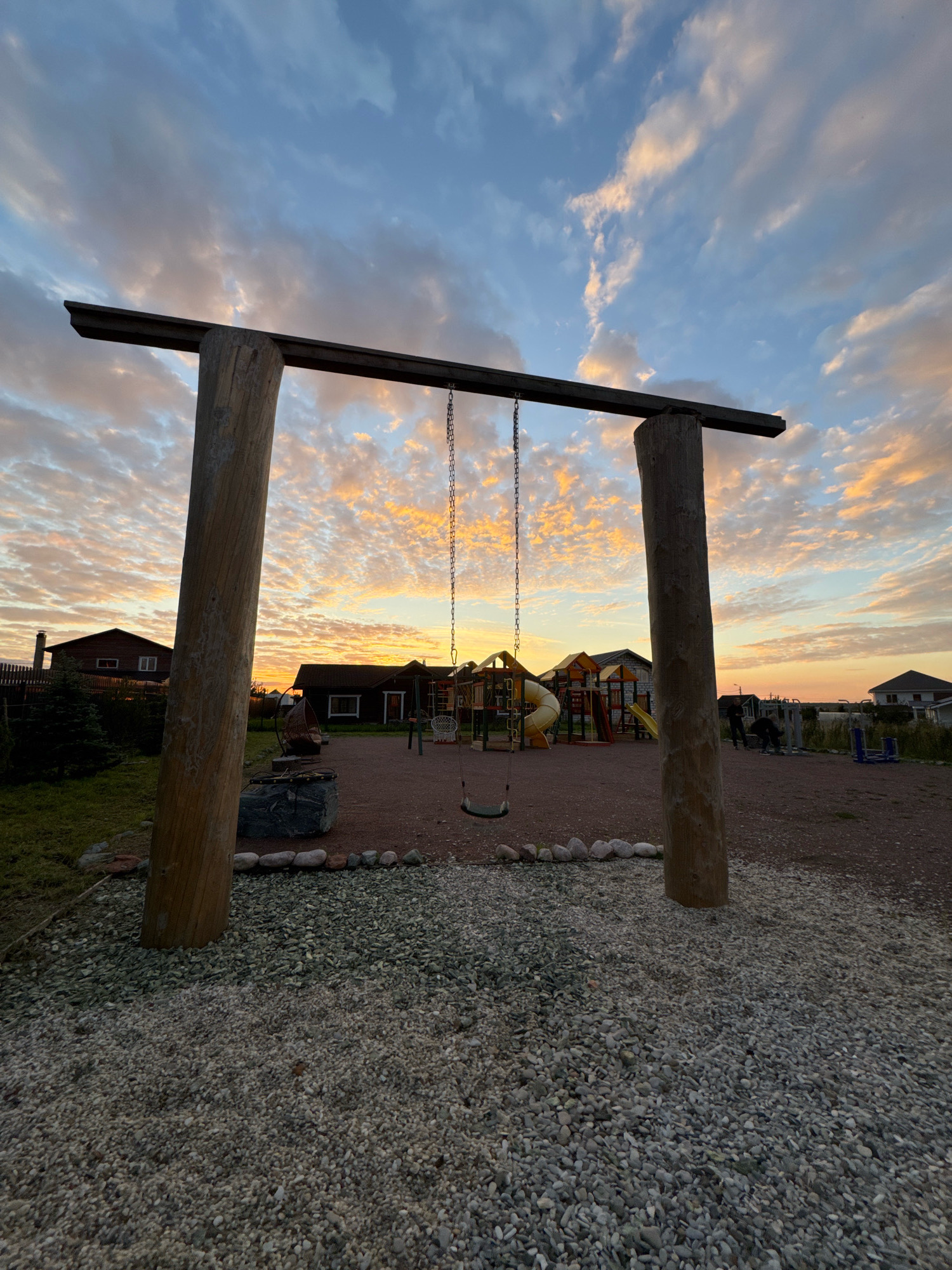 The image shows a playground at dusk in Timmolovo, Russia. A wooden swing set stands in the foreground, with a single swing hanging from its chains. In the background, a playground structure with slides and climbing elements is visible, along with several residential buildings. The sky is filled with dramatic clouds illuminated by the setting sun, casting a warm orange and yellow glow. The ground is covered in gravel. Two individuals are seen in the distance, near the playground equipment.