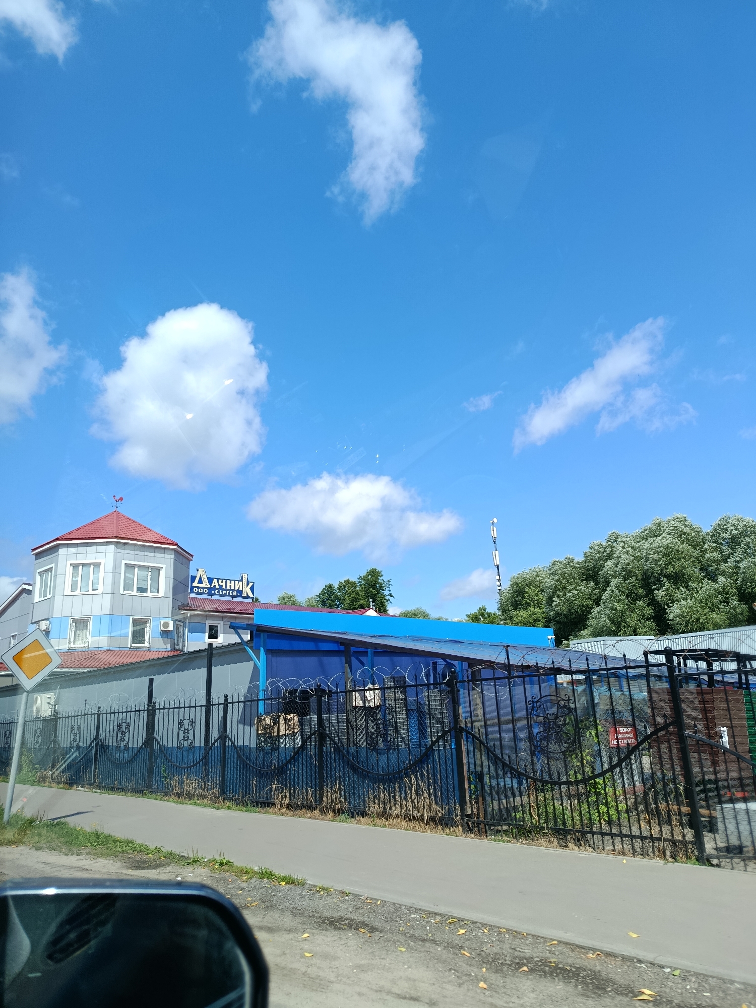 A street-level view of a commercial building in Orekhovo-Zuyevo, Russia, features a light blue sky with scattered white clouds. The building, with a red-tiled octagonal tower topped by a weather vane, is gray and light blue with several windows. A sign with blue lettering that reads "ДАЧНИК" is visible above an adjacent structure with a blue roof. A black metal fence with barbed wire runs along the front of the property, separating it from a paved road and a sidewalk. A yellow diamond-shaped road sign is positioned near the left edge of the frame. In the background, a tall communication tower stands against the horizon, partially obscured by dense green trees. The reflection of a car's rearview mirror is visible in the lower left corner of the image.