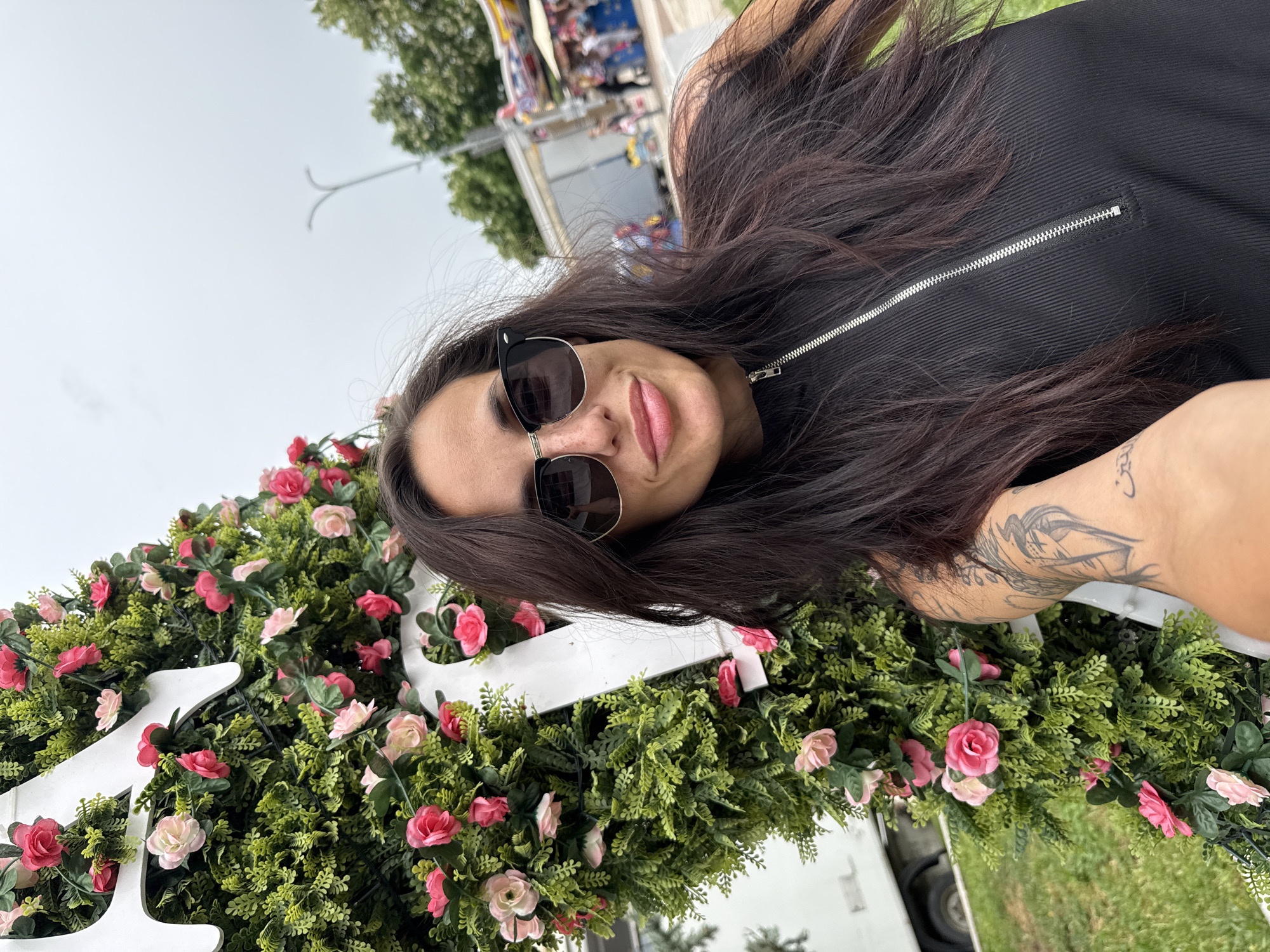 A woman with long brown hair, sunglasses, and a tattoo on her arm is posing for a photo. She is wearing a black zippered top. In front of her is a floral arrangement of greenery and pink roses, set against white lettering. The background includes a grassy area, trees, and a glimpse of structures that appear to be part of a fair or festival. The scene is outdoors and appears to be in Mezdra, Bulgaria.