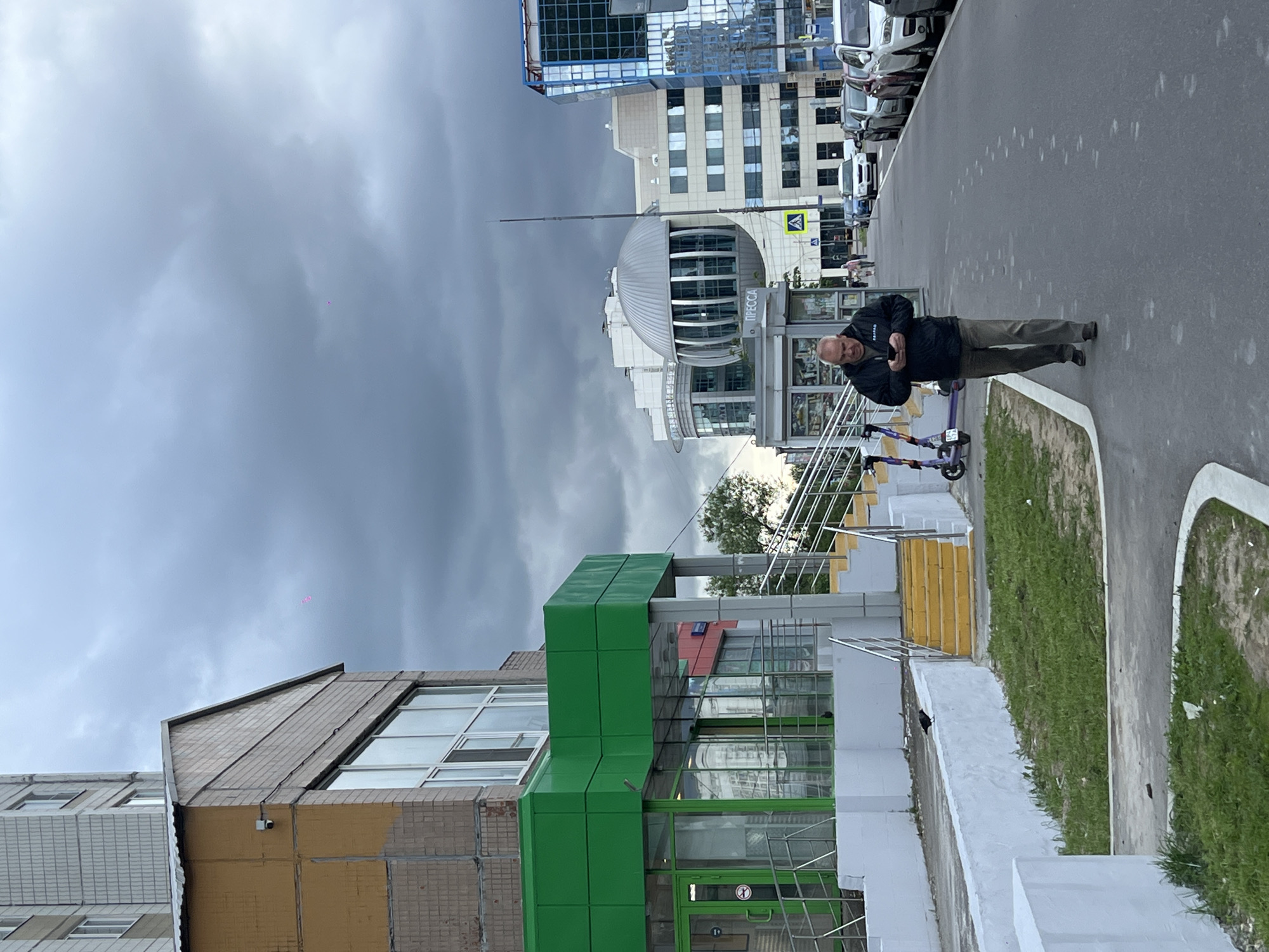 The image shows a man standing on a sidewalk in what appears to be Moscow, Russia. He is holding a smartphone and is dressed in a dark jacket and pants. A purple scooter is next to him. Behind him is a street and several buildings, including one with a green facade and a building with a sign that says "Пресca" (Press). The sky is overcast.