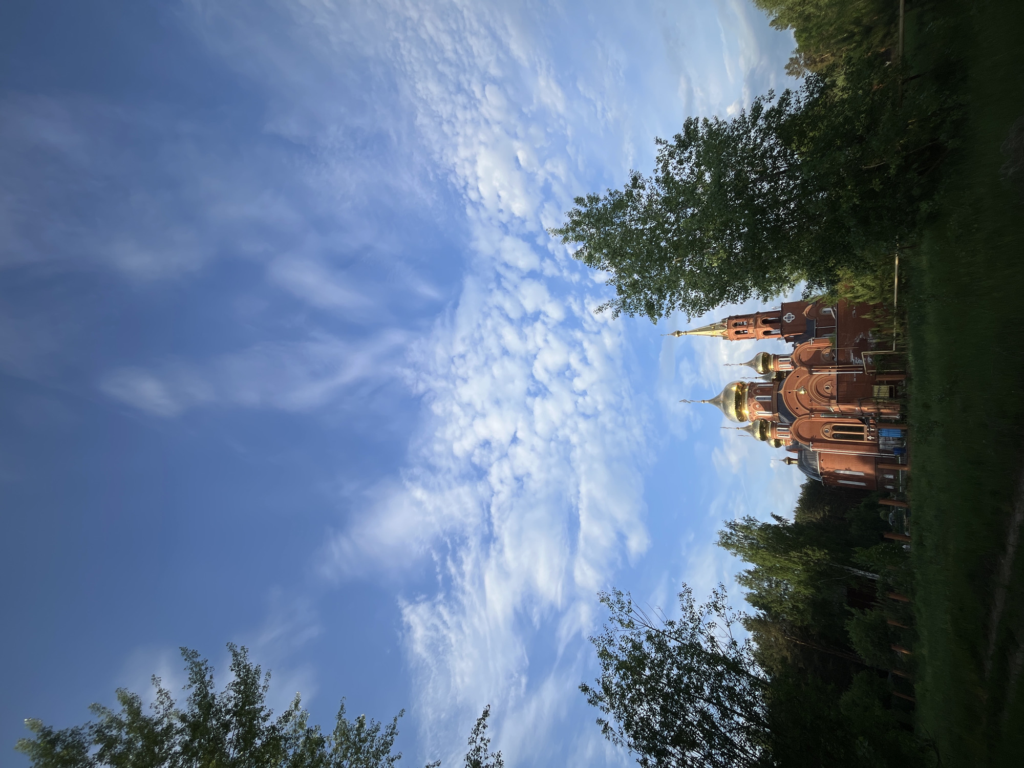 The image shows a brick building with gold domes, likely a Russian Orthodox church, positioned between trees and under a blue sky with streaky clouds. The building is ornate with multiple levels and a spire. The scene is in the city of Chaikovsky, Russia, judging from the architecture. There is a park or grassy area to the side of the building. The picture is oriented vertically, possibly taken by a phone. No people are visible in the image.