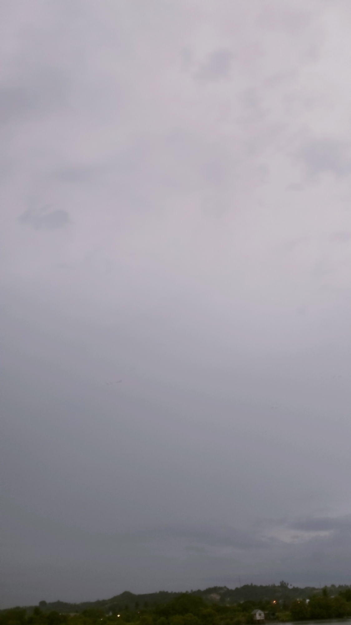 The image captures a serene yet somber landscape dominated by a vast, overcast sky. The sky, which occupies the majority of the frame, is a uniform dull gray, indicating heavy cloud cover or an impending storm. Subtle variations in cloud density are visible, with slightly darker patches suggesting deeper formations.
In the lower portion of the image, a narrow strip of land extends across the horizon. This land is covered with dense, dark green foliage, indicative of abundant trees and vegetation. A calm body of water is visible in the foreground, stretching along the base of the land, reflecting the muted light.
Towards the right side of the frame, near the water's edge and nestled among the trees, a small, solitary white structure with a dark roof can be discerned. This appears to be a small house or shed, adding a touch of human presence to the natural scene. Further inland, barely visible through the vegetation, are a few faint, yellowish light sources, possibly from distant buildings or streetlights.
The overall atmosphere suggests a heavily overcast day, possibly just before rain, during dusk, or early morning. There are no people visible, and no specific activity is taking place. The scene conveys a sense of quietude and the calm before or after a weather event. There is no visible text in the image. The location context of Lhokseumawe, Indonesia, aligns with the lush tropical vegetation and coastal/riverside environment depicted.