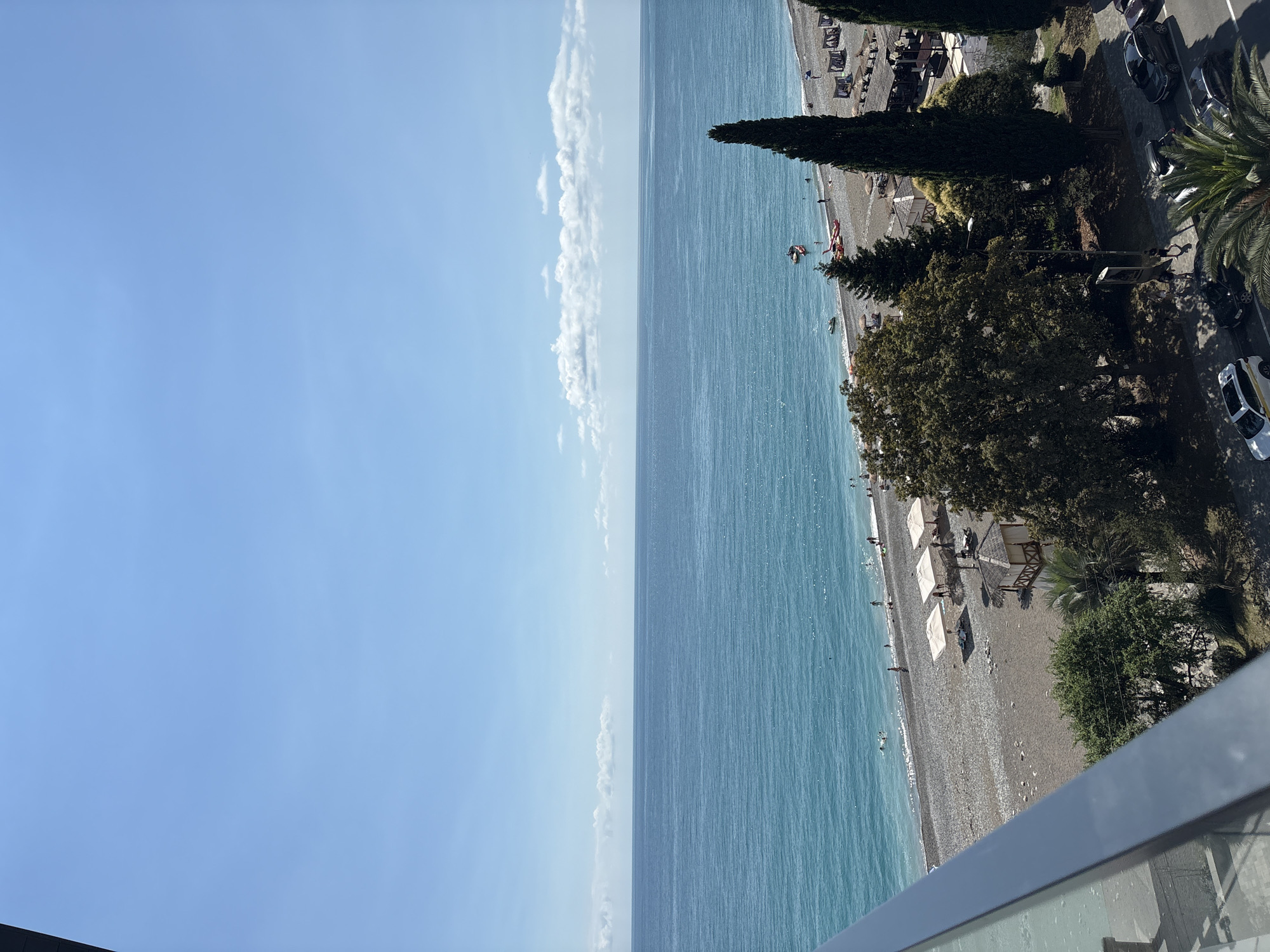 The image captures a coastal scene in Abaata, Abkhazia, under a clear blue sky with sparse white clouds. The foreground shows a balcony railing. Below, a pebble beach stretches along the turquoise sea, populated by a few beachgoers. Several umbrellas are set up on the sand, and a small, wooden lifeguard or changing station is visible. Adjacent to the beach is a strip of greenery with trees, including tall cypress trees, and some buildings with covered seating areas. A paved road runs parallel to the greenery, with several cars parked along it. People are seen walking on the sidewalk and on the beach. The overall impression is of a sunny day at a seaside resort.
