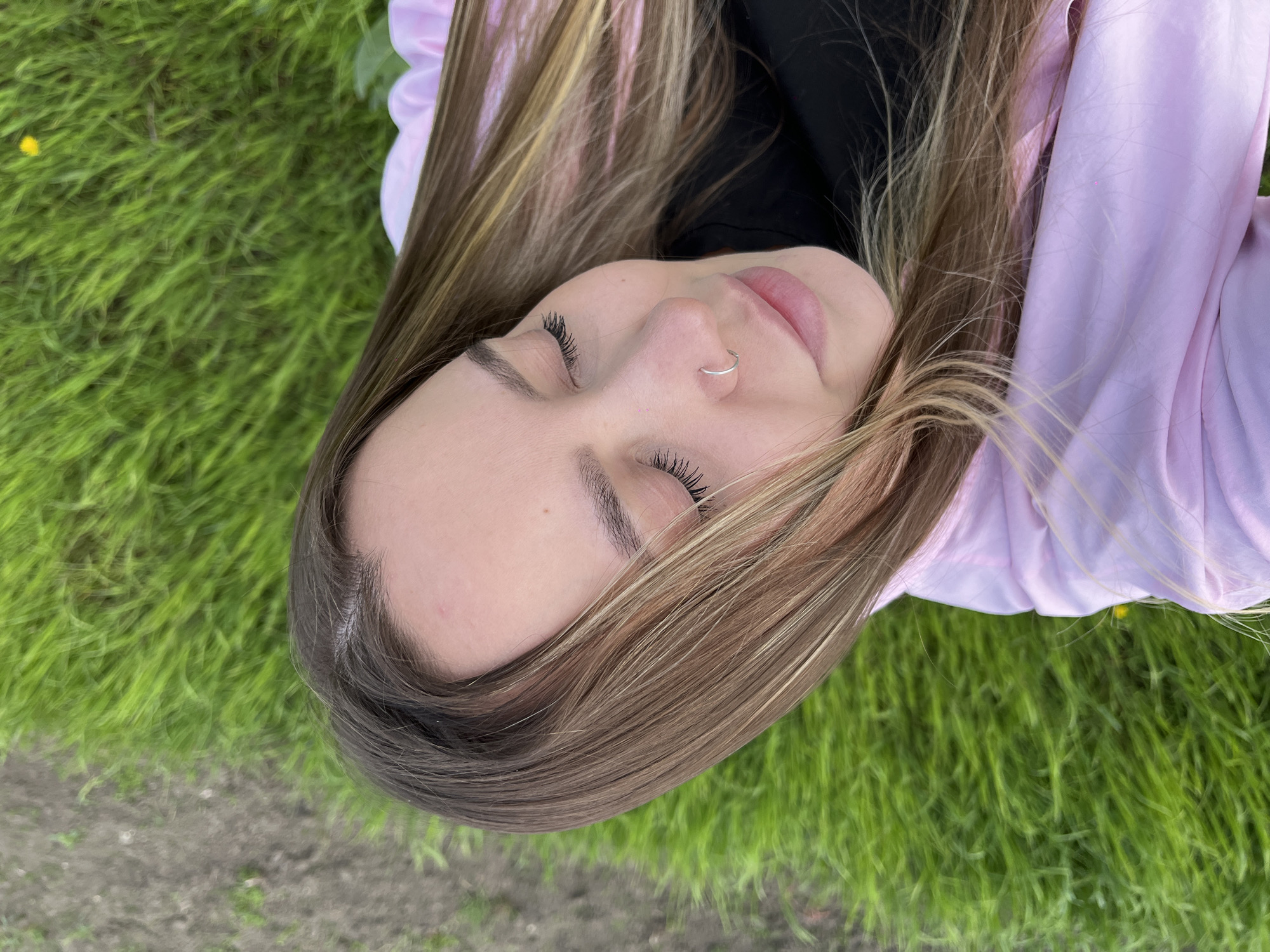 The original image depicts a close-up, high-angle view of a young woman lying peacefully on vibrant green grass. Her eyes are closed, revealing long, dark eyelashes, and her lips are slightly parted. A small, simple silver hoop nose ring is visible in her left nostril. She has long, straight hair, streaked with shades of light brown and blonde, which spreads across the grass and a light pink jacket or top she is wearing, along with a black inner layer.
The setting is outdoors, dominated by the lush green grass, which suggests a natural environment like a park or field. A small yellow dandelion flower can be seen in the grass towards the upper left, and some patches of darker soil are visible in the lower part of the frame. The lighting appears soft and even, indicating a serene and tranquil atmosphere, possibly on an overcast day. The overall mood conveyed is one of relaxation and calm, potentially in Novopokrovskaya, Russia. There is no visible text in the image.