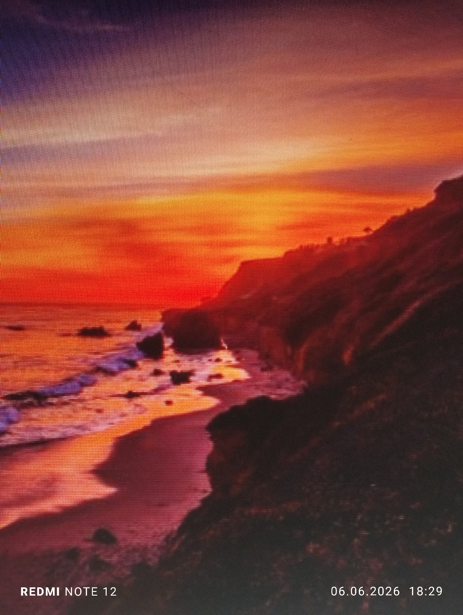 The image depicts a vivid coastal sunset in the city of Burgas, Bulgaria, captured as a photograph of a digital screen. The sky fills the upper portion of the frame, displaying a dramatic gradient of colors, transitioning from deep indigo and purple at the top, through warm oranges and yellows, to an intense red along the horizon. Streaky, layered cloud formations are visible, reflecting these vibrant hues.
On the right side, a rugged coastline is featured, comprising dark, silhouetted cliffs and hills that descend towards the water. Subtle outlines of sparse vegetation, consistent with trees or shrubs, are discernible on the higher elevations. To the left, the ocean meets a beach, appearing to be sandy or rocky. Waves are observed breaking along the shoreline, reflecting the bright red and orange light from the sky, creating streaks of purple and white foam. Several dark rock formations are scattered both within the water near the shore and directly on the beach.
No people, specific man-made structures, or direct interactions are visible within the scene.
The photograph exhibits a visible grid pattern across its surface, indicative of being a picture taken of a digital display. Embedded text at the bottom left identifies "REDMI NOTE 12," while a timestamp at the bottom right indicates "06.06.2026 18:29."