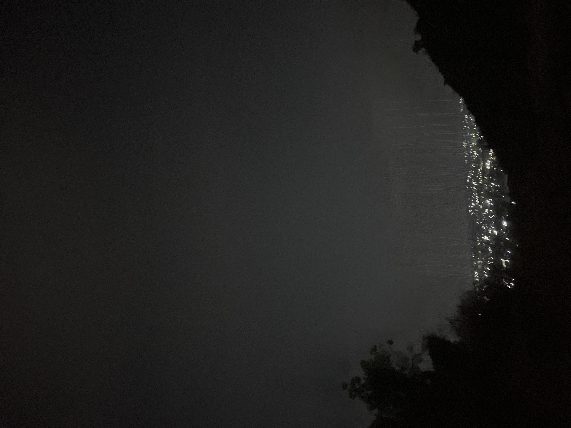 The image appears to be a nighttime view of a city from an elevated position, likely a hill or mountain. The city lights are visible as bright points against a dark background. There is a rock formation or cliff in the foreground, partly obscuring the view of the city. The dark, blurry areas at the bottom of the image suggest trees or vegetation. The streak-like lights may be caused by a camera movement or a long exposure setting. The location is indicated to be Jos, Nigeria.