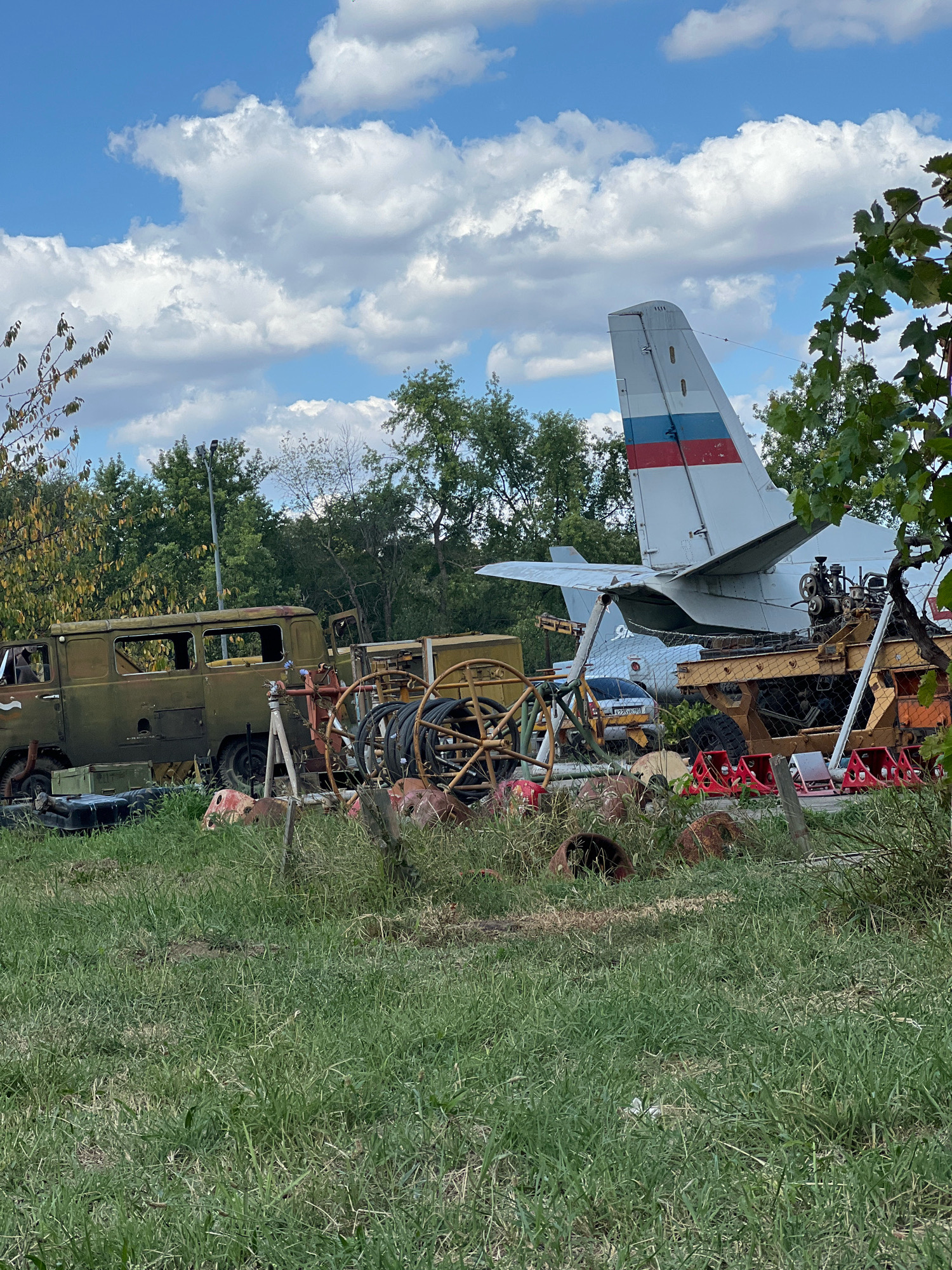 The image depicts an outdoor scene with various weathered vehicles and equipment situated in a grassy area, with trees and a cloudy sky in the background. The central focus is the tail section of a white airplane with a red, white, and blue stripe pattern, indicative of Russian national colors. To the left, a faded olive-drab military-style truck, likely a UAZ-452, is visible, appearing derelict with open doors and windows. In front of the airplane and truck, a collection of rusty metal objects, including what appear to be spools of cable or hose, machinery parts, and a yellow apparatus, are scattered amongst the tall grass. A chain links some of these objects together. Further to the right, the rear end of another vehicle or piece of equipment, painted yellow, is partially obscured by foliage. Red and white traffic barriers are also present. The environment appears to be an open field or storage area, possibly a junkyard or an abandoned airfield. No people are visible in the image. The setting is identified as the city of Belozernyi, Russia.