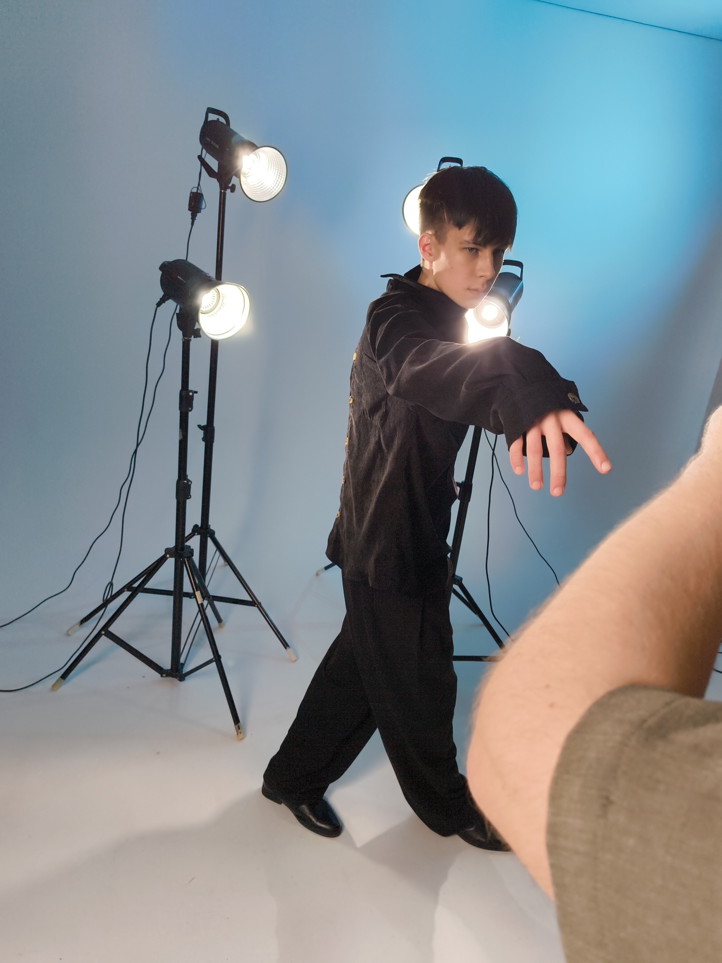 A photograph shows a person in a black suit striking a pose in a photography studio setting in the city of Chaikovsky, Russia. Two light stands with halogen lights are positioned on the left and right sides of the subject. The subject's hair is styled short and dark. The studio background and floor are light blue. The person is wearing black leather dress shoes. An arm belonging to a photographer is partially visible in the lower right corner of the frame, holding a camera.