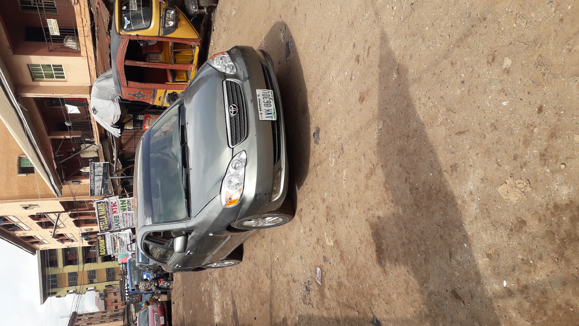 The image depicts a street scene in Onitsha, Nigeria. A grey Toyota Corolla is parked in the foreground, facing the camera. The car has a license plate reading "AWK-863DU" and appears to have some minor cosmetic damage on its front bumper. Behind the car, a yellow and orange auto-rickshaw is visible, partially obscured by the car. The street is unpaved, covered in dirt and gravel, suggesting it may be a less developed area of the city. Buildings line the street, some appearing to be residential or commercial establishments with signs hanging from them. One sign reads "STELLA MARIS" and another, partially visible, mentions "CHIDMA RESTAURANT". The overall impression is that of a busy, typical urban environment in Nigeria, with a mix of vehicles and pedestrian activity implied by the surrounding infrastructure.