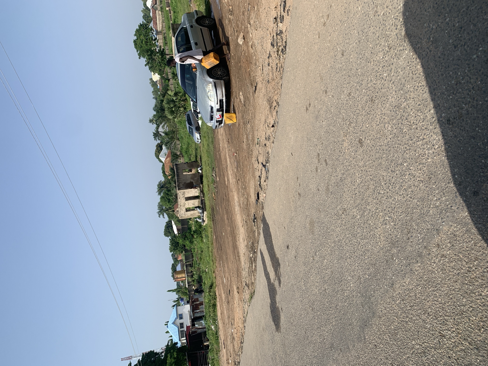 A man washes a silver sedan on a dirt lot next to a paved road. The car is parked in front of a partially constructed building. In the background, there are several houses and trees. The sky is clear and blue.