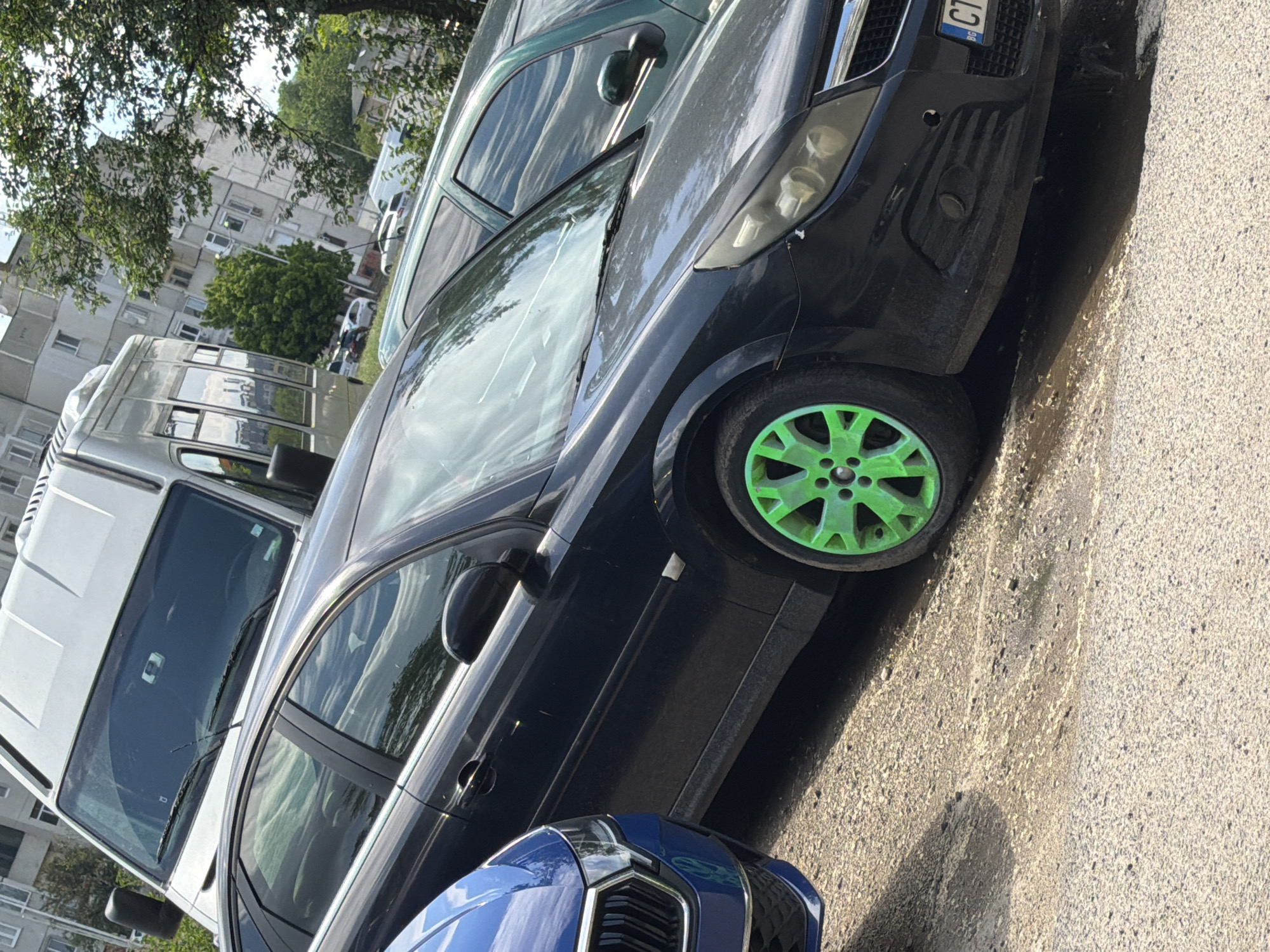 The image depicts a street scene in Stara Zagora, Bulgaria. A dark-colored sedan with bright green rims is parked. Part of a blue car is visible in the foreground. A white van is positioned to the left of the sedan. The sedan displays a Bulgarian license plate, the letters "CT" being visible. Buildings and trees are present in the background. The image appears to be taken during daylight.