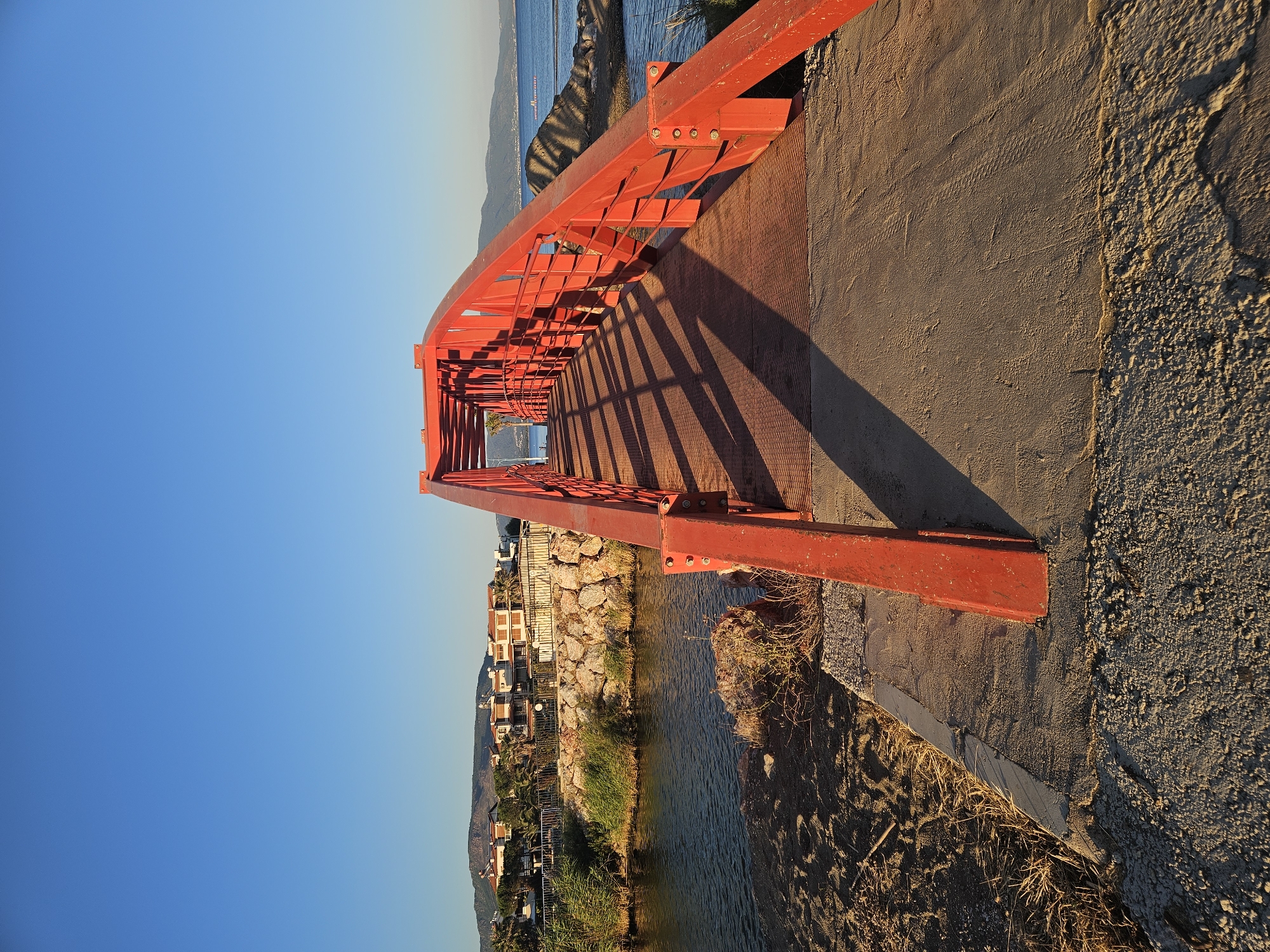 A bright red pedestrian bridge spans across a narrow body of water, casting long shadows on its metal walkway. The bridge is constructed with a lattice-like railing and appears to be made of painted metal. On the left bank of the water, there is sandy ground with some dry grass and reeds. Further back from the water's edge, a low stone wall separates the bank from a grassy area and several houses with light-colored walls and tiled roofs, set against a backdrop of green trees and hills. On the right side of the bridge, the water extends towards the sea, with a rocky shoreline and more distant hills visible under a clear blue sky. The scene suggests a coastal or riverside location, likely in a town or residential area. This bridge is located in the Akkum neighborhood of Seferihisar, Izmir, Turkey, specifically crossing the narrow channel connecting the Akkum lake to the Aegean Sea.