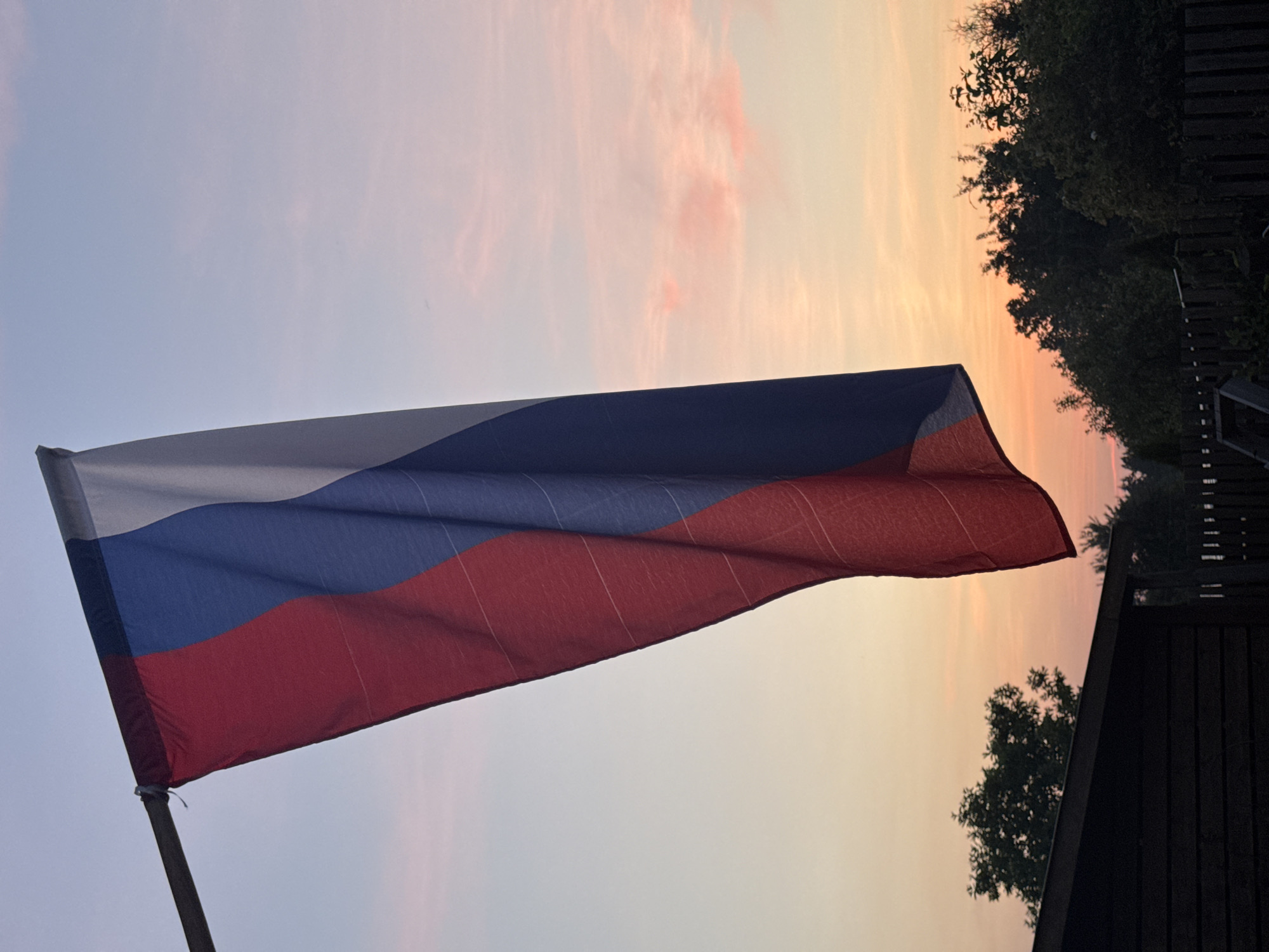 The image depicts the Russian flag hanging from a flagpole against a sky at dusk. The flag is predominantly red, blue, and white, arranged in vertical stripes. The sky exhibits a gradient of colors, transitioning from a pale blue at the top to soft oranges and pinks near the horizon, suggesting either sunrise or sunset. In the background, silhouetted trees and a dark, low-lying structure, possibly a shed or fence, are visible. The scene appears to be an outdoor setting, likely a residential or rural area, with no people present. The overall atmosphere is serene and quiet.