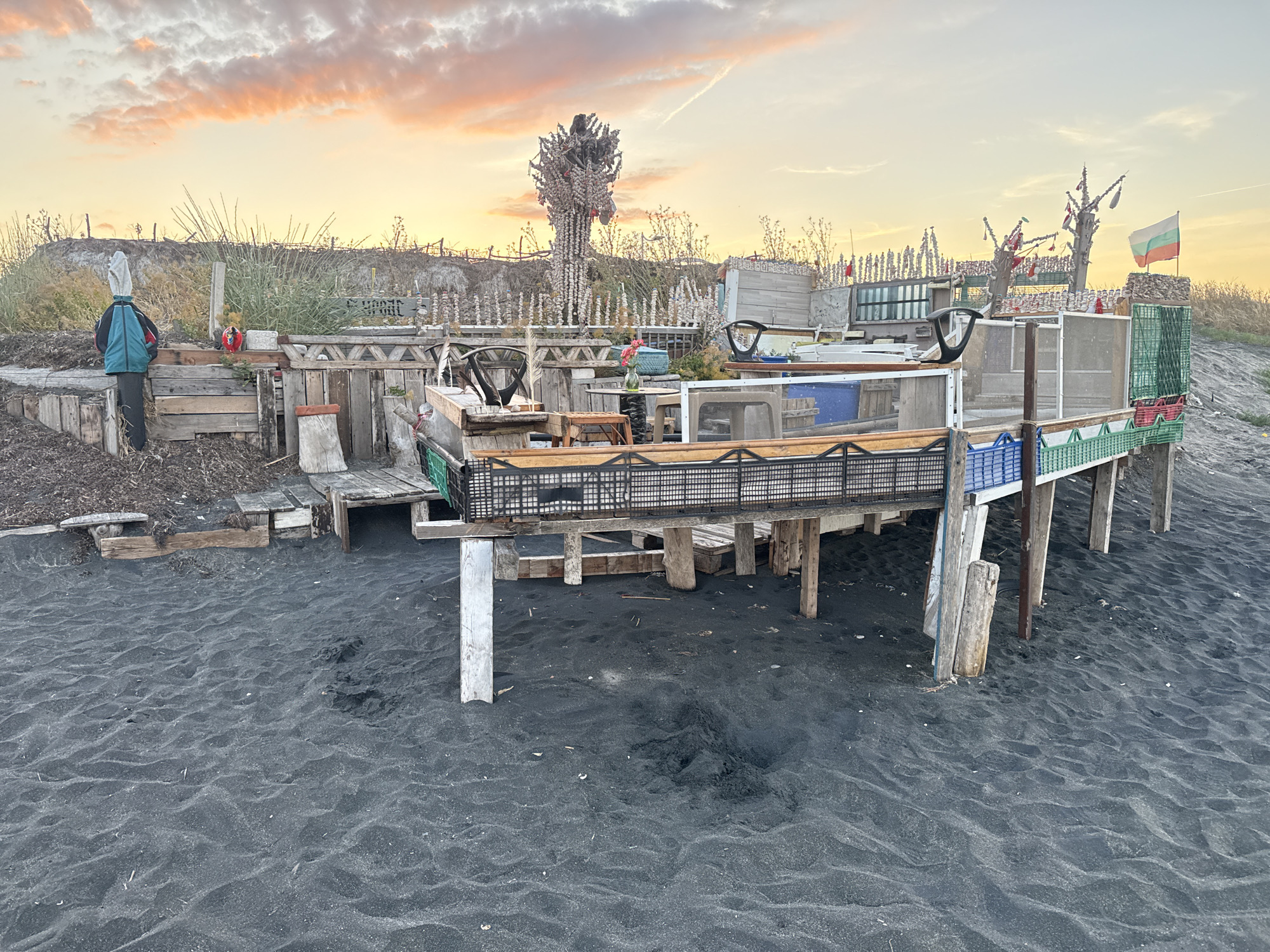The image depicts an outdoor, possibly makeshift, structure situated on a dark sandy beach, likely in Burgas, Bulgaria. The structure appears to be constructed from salvaged materials, including wood planks, metal grates, and plastic crates, forming platforms and enclosures. A scarecrow-like figure, wearing a blue and green backpack, stands to the left of the structure. Further to the right, a Bulgarian flag is visible, attached to a tall pole, flying against a sky with soft, orange and pink clouds, suggesting sunset or sunrise. The overall scene is rustic and appears to be an unconventional beach installation.