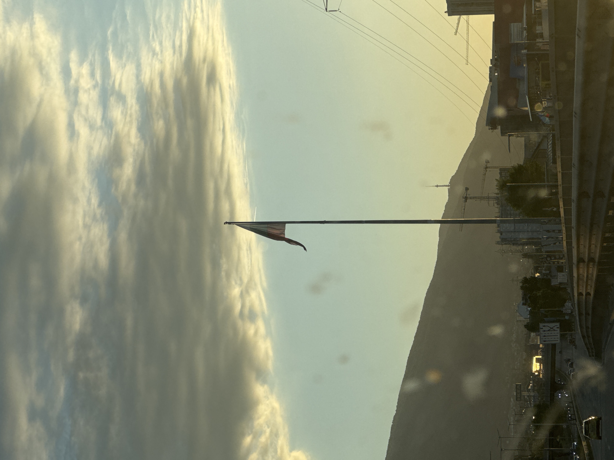 The image shows a flag waving in the wind against a backdrop of a cloudy sky and a mountain range. There are power lines and construction cranes visible in the distance. A city with buildings and a road is also present. The location is likely Sofia, Bulgaria, given the presence of Vitosha Mountain. The image appears to have been taken from inside a vehicle due to the presence of glare on the window.