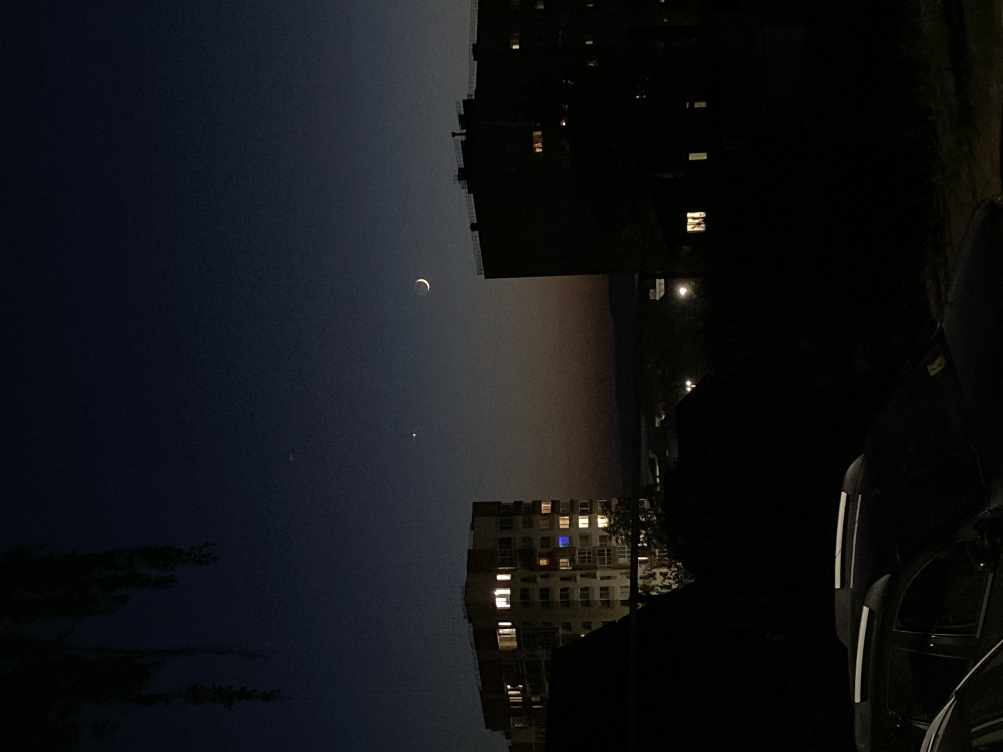 The image depicts a night scene in Votkinsk, Russia. A crescent moon is visible in the upper left quadrant of the sky, positioned above a single bright star or planet. Below them, the sky transitions from dark blue to a gradient of orange and pink, suggesting either sunrise or sunset.
Two buildings dominate the lower part of the frame, both displaying lit windows indicating occupancy. One building is relatively close to the viewer and appears as a dark silhouette, while the other is further away and shows more detail, including individual windows with light emitting from them.
In the foreground to the right, parts of dark-colored vehicles are visible, indicating a parked or moving car, with some details like a stripe from a tail light slightly visible. The environment is dark with minimal lighting. The image seems to be taken from ground level, providing a view of the sky, buildings, and nearby parked vehicles.