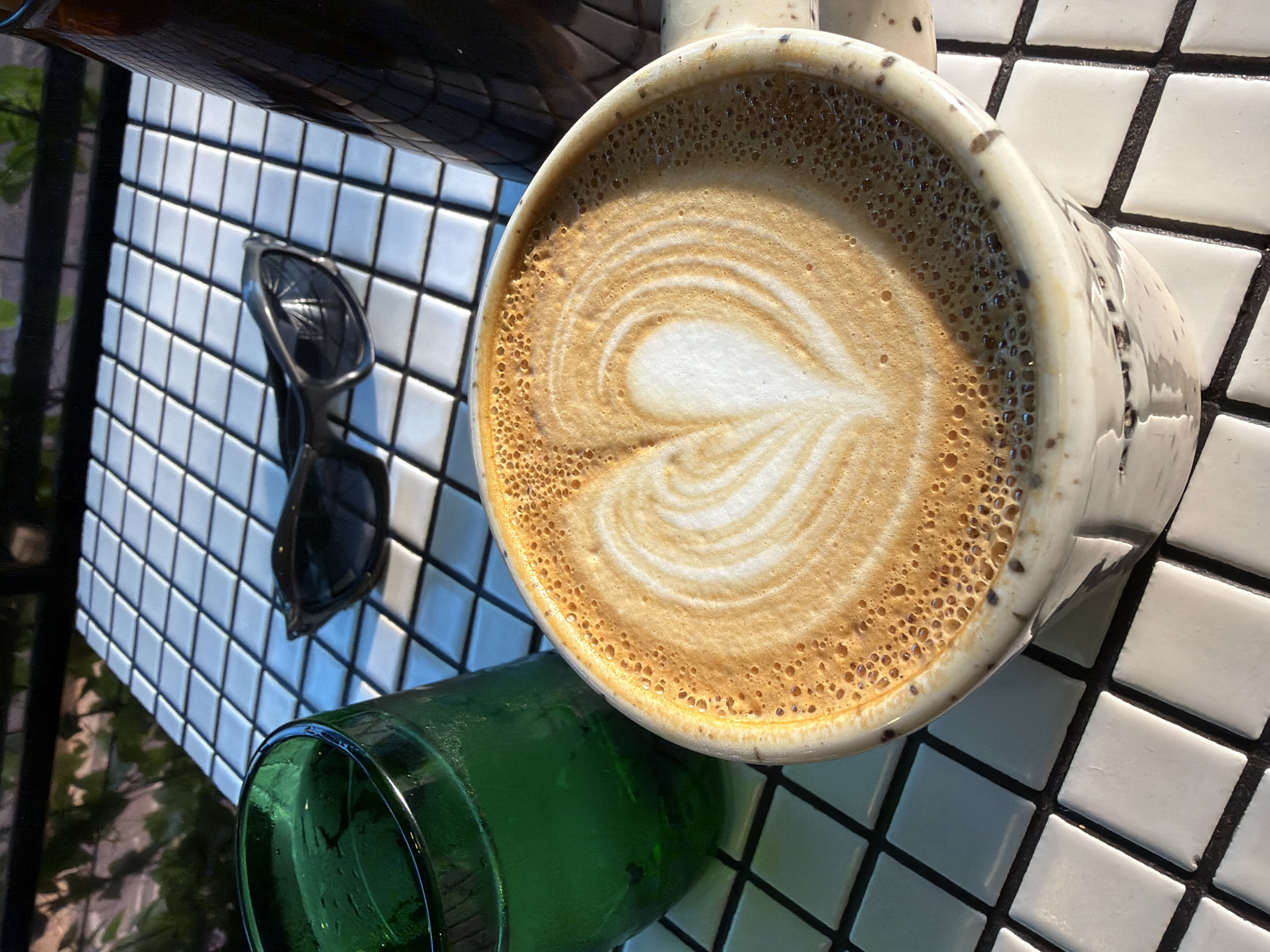 The image shows a top-down view of a table with white and black square tiles, possibly in a café or outdoor setting. On the table is a mug of coffee with latte art in the shape of a heart, a green glass, and a pair of black sunglasses. The location is possibly in North Kuta, Indonesia. The lighting suggests it is daytime.