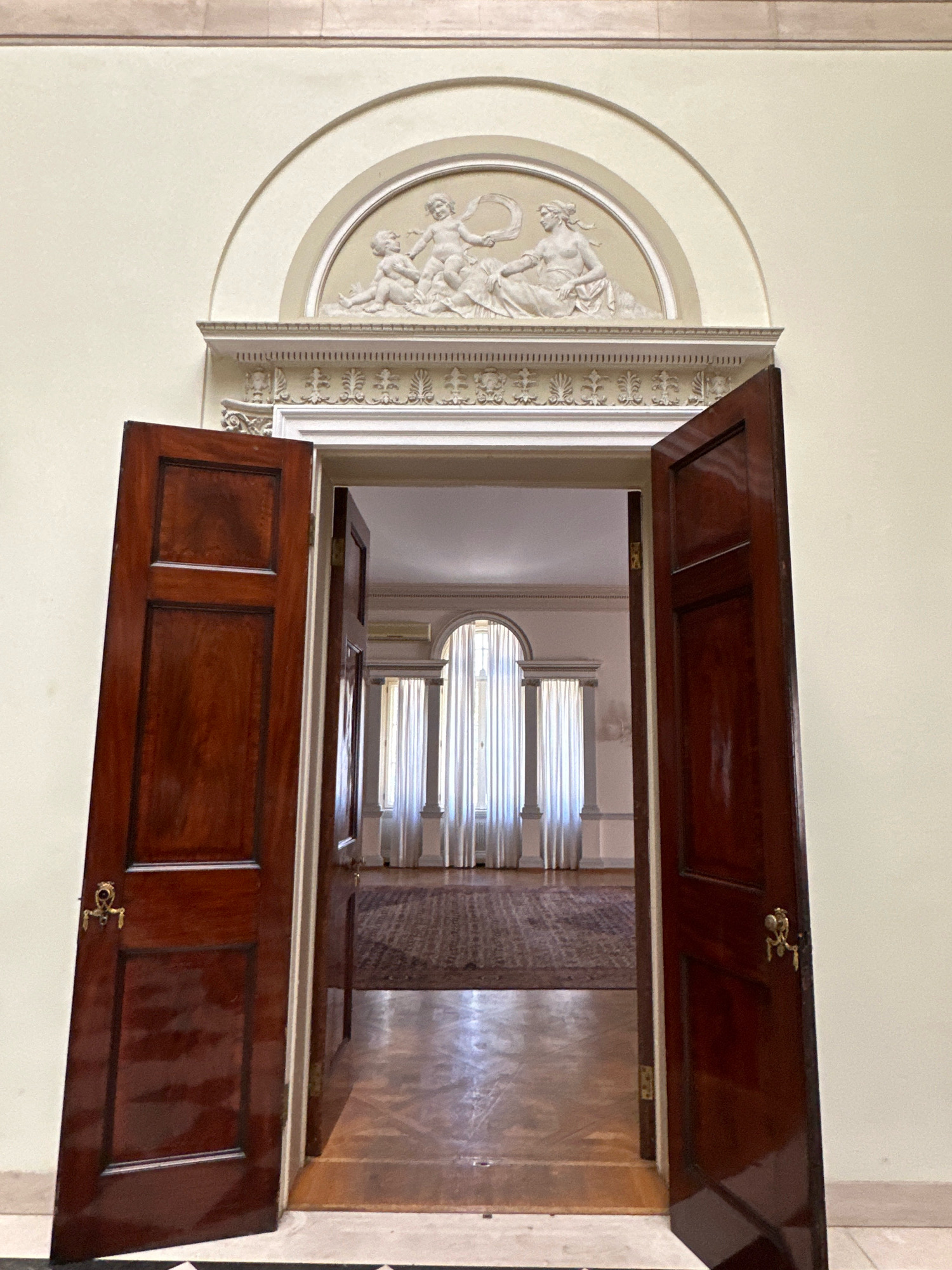 The image depicts an interior view of a grand doorway in Belgrade, Serbia, likely within a historical building or museum such as the National Museum of Serbia. The doorway features two imposing dark wood doors, both ajar. The left door is angled inward, revealing its paneled surface and a decorative brass handle. The right door is also open, leading into a well-lit room beyond.
Above the doorway, an arched architectural detail frames a bas-relief sculpture. This sculpture appears to depict mythological figures, possibly cherubs or putti, interacting around a circular object, rendered in a classical style. Below the arch, a decorative frieze with intricate patterns runs across the wall.
The room visible through the doorway is spacious, with a patterned rug covering a significant portion of the wooden floor. Tall, draped curtains frame large arched windows at the far end of the room, allowing natural light to illuminate the space. The overall impression is one of elegance and historical significance.