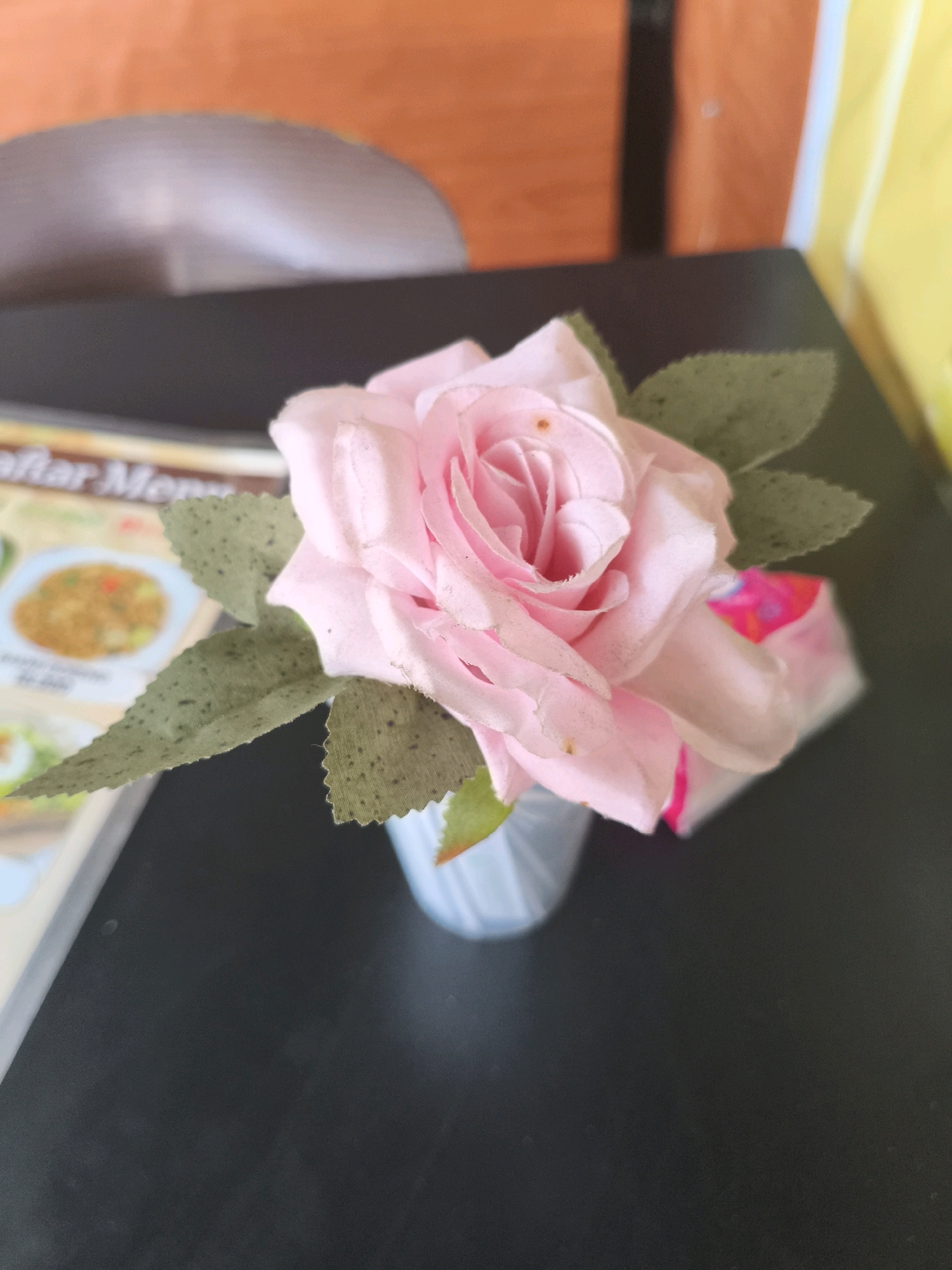 A light pink artificial rose is displayed in a small, light blue vase on a dark, reflective surface, possibly a table in an eatery or cafe in Depok, Indonesia. The rose, with visible green leaves that appear somewhat weathered, is the primary focus. To the left of the rose, a blurred menu or pamphlet featuring an image of food is partially visible. The background is out of focus, showing areas of orange and yellow, suggesting a wall or partition. A small, bright pink and white object, possibly packaging, is situated behind the rose and slightly to the right. The lighting suggests an indoor environment.