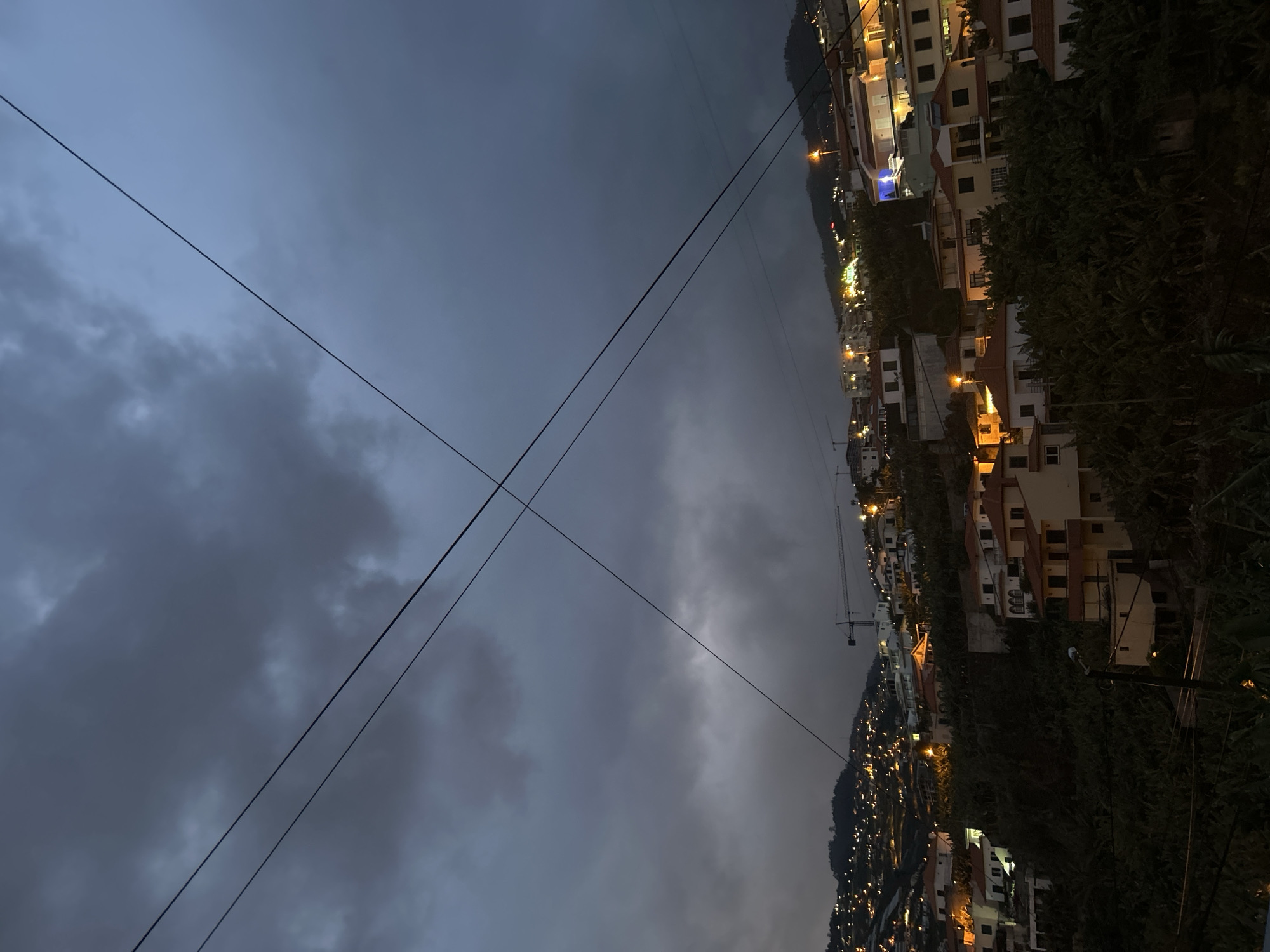 The image shows a hillside view in Câmara de Lobos, Portugal, at night or dusk. Illuminated buildings, primarily residential, are nestled on the incline. The sky is overcast with heavy, dark clouds. Power lines intersect diagonally across the view. Vegetation, likely trees and shrubs, are visible on the right side of the frame. A crane is visible in the distance.