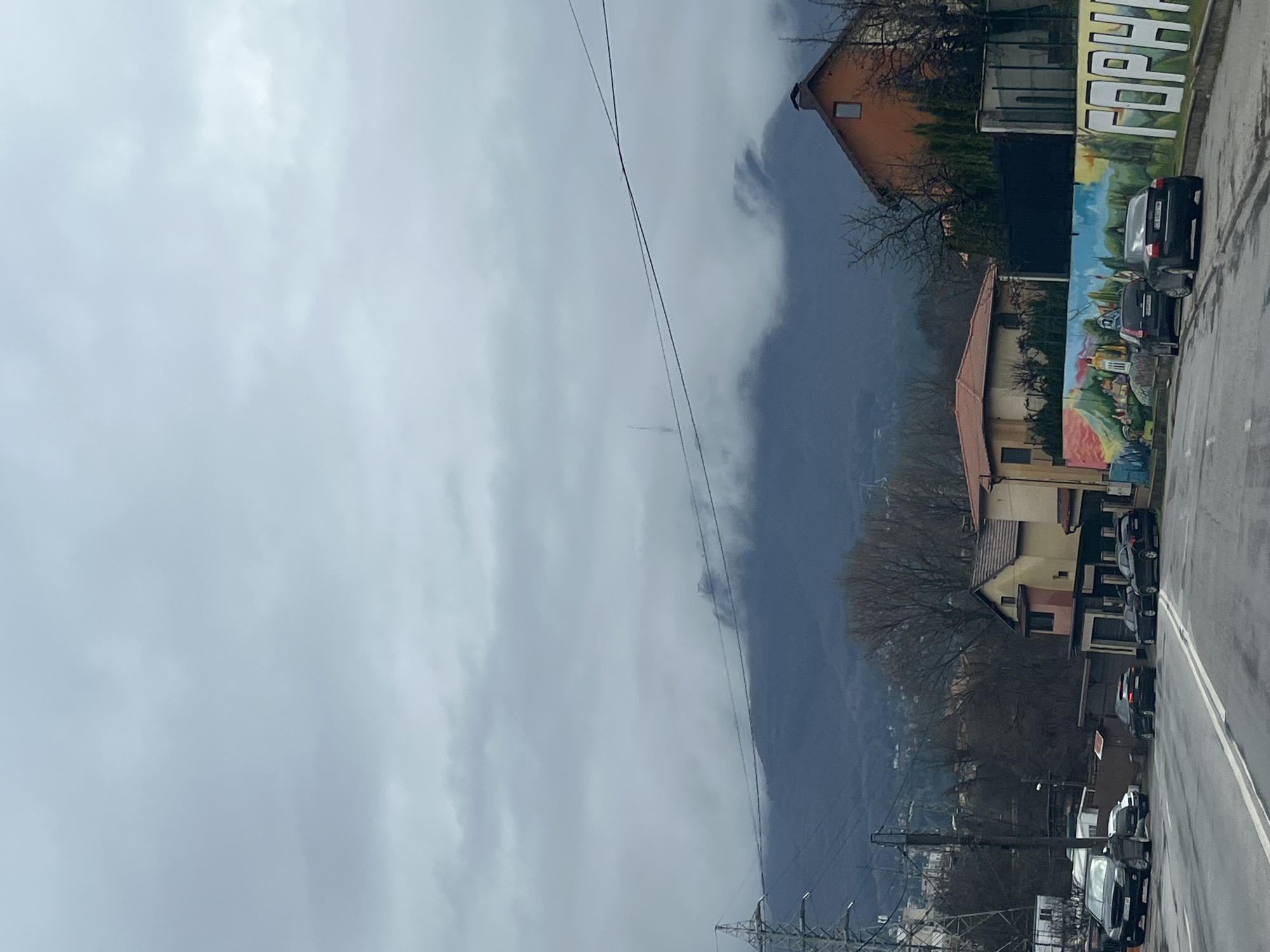 The image shows a street scene with buildings and parked cars on the right side, with a mountain in the background obscured by heavy clouds. Electrical power lines are visible in the foreground. A mural is displayed on the side of a building, with some letters in English and Bulgarian. The location is possibly in the vicinity of Sofia, Bulgaria, considering the architectural styles and the presence of Cyrillic text.