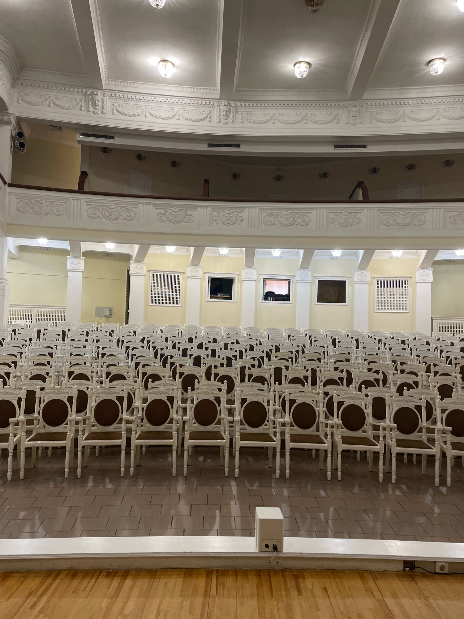 The image depicts the interior of a large, ornate hall, likely a theater or concert hall, filled with rows of empty white chairs with brown seats. The hall features high ceilings with decorative moldings and chandeliers. Several windows, some with grates, are visible on the walls, along with what appear to be ventilation grilles. The chairs are arranged on a tiled floor, and in the foreground, a stage or raised platform is visible, with a small white speaker box and electrical outlets. The overall impression is one of a formal, unoccupied space. The image was taken in the Omsk State Philharmonic Hall in Omsk, Russia.