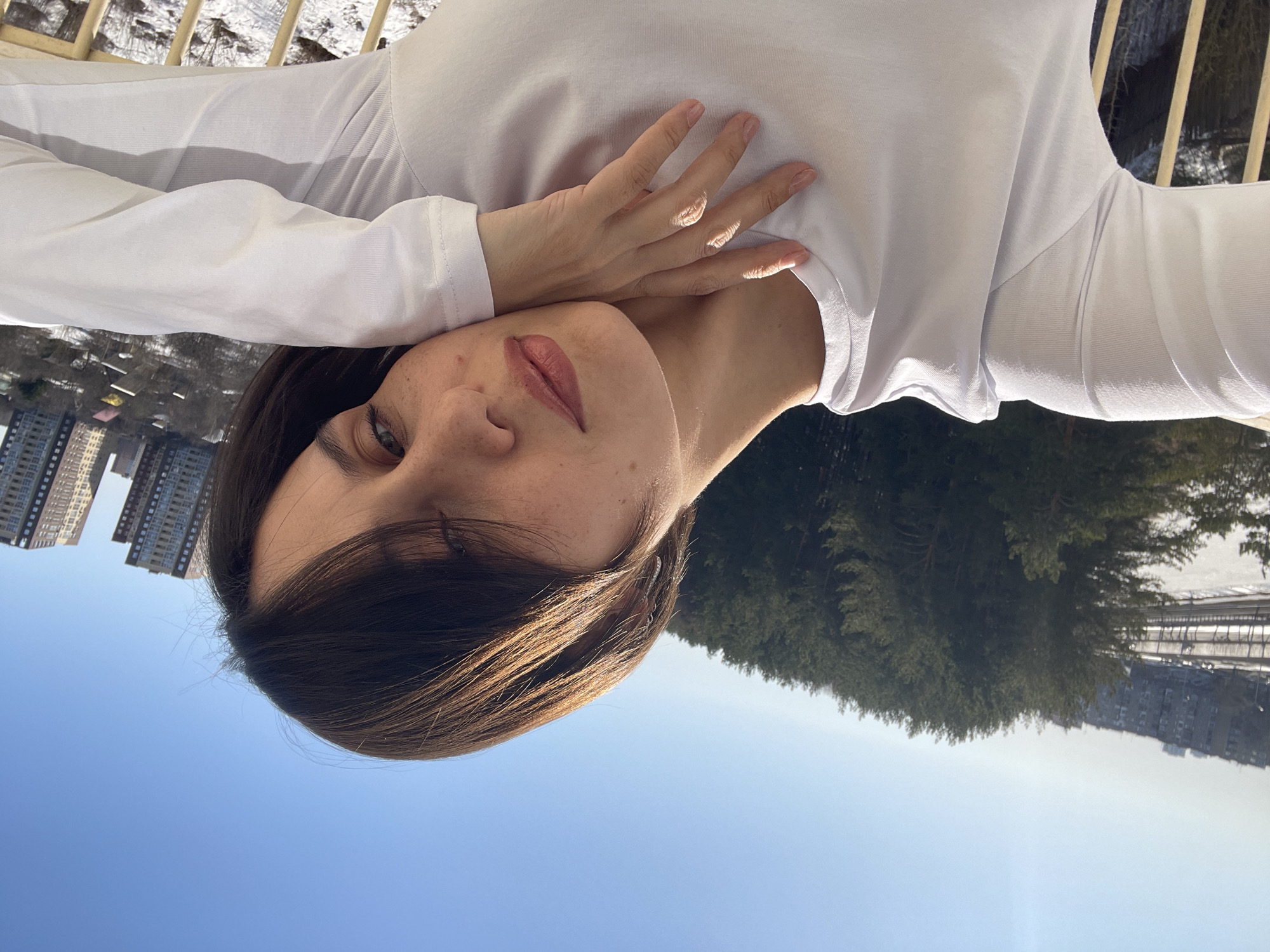 A young woman with long brown hair and freckles is lying on her back, wearing a white long-sleeved shirt. Her right hand is resting on her chest. Behind her is a blue sky, and various scenes of Izhevsk, Russia are visible. To the left are tall apartment buildings. To the right, there is a dense forest and a snow-covered area with a fence. The image is oriented so that the woman appears to be lying on the sky.