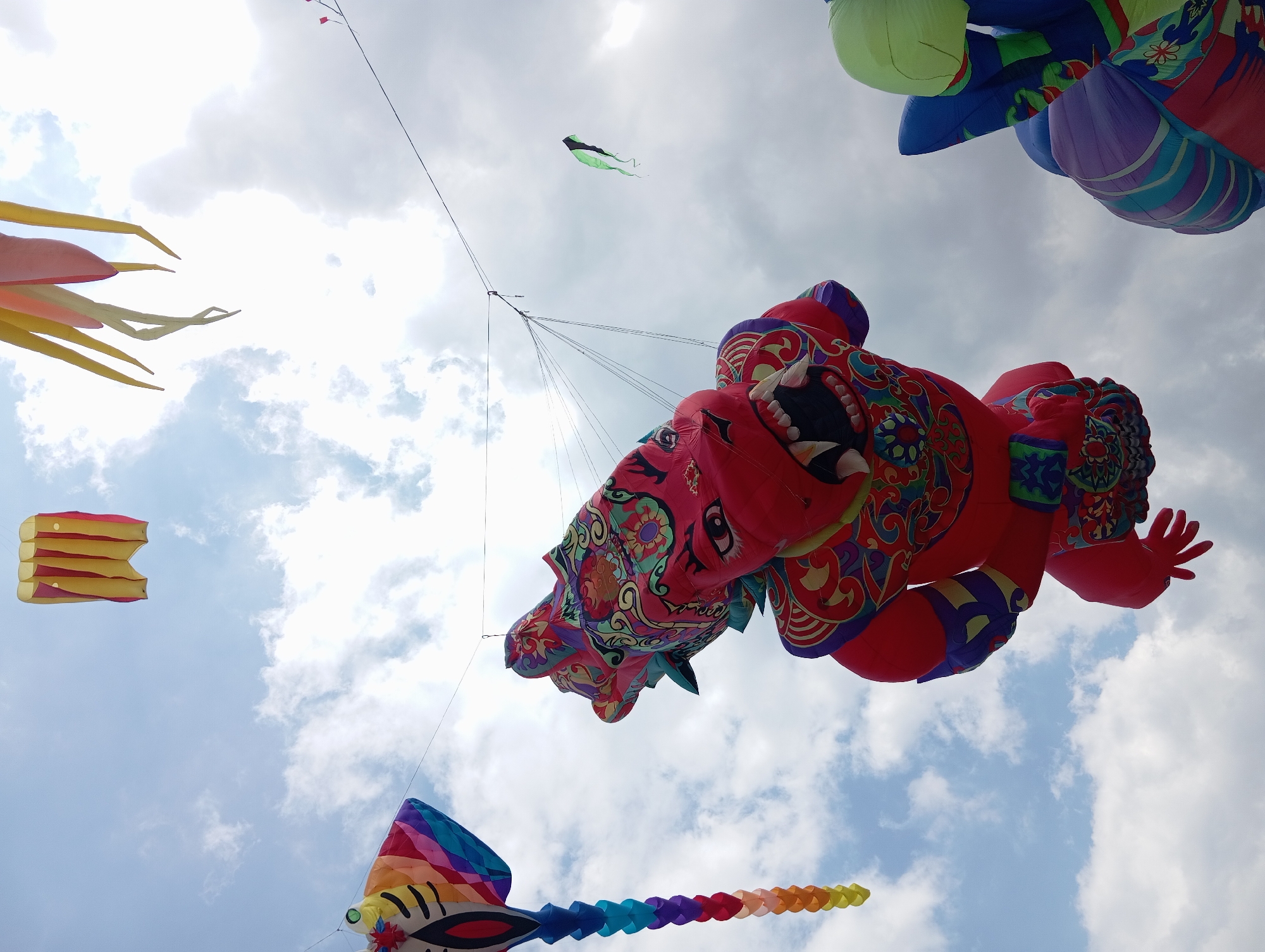 The image depicts a collection of colorful kites flying in a cloudy sky, likely at a kite festival. In the foreground, a large, red kite shaped like a mythical creature with sharp teeth and ornate, colorful patterns dominates the center of the frame. To the left, a kite with a rainbow-colored tail hangs vertically. In the background, other kites are visible, including a small green and black kite and a yellow and red rectangular kite. The scene is taken from a low angle, looking up at the kites against the sky. The location is identified as Parangtritis, Indonesia, suggesting a coastal setting often associated with kite flying due to consistent winds. No people are visible in the frame.