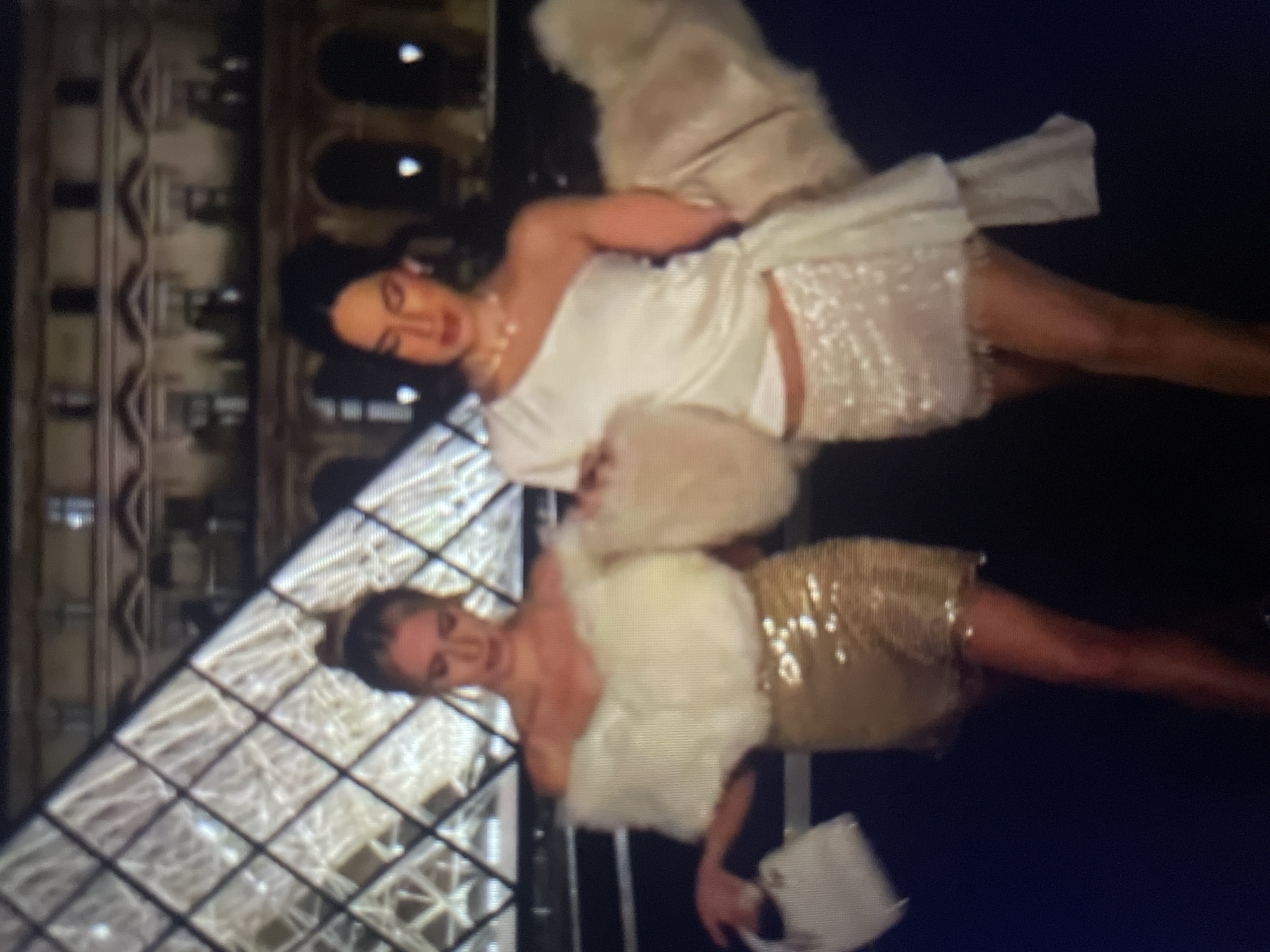 Two young women are standing in front of a glass pyramid and a building with arched windows. The woman on the left has blonde hair pulled back and is wearing an off-the-shoulder white top with fluffy trim and a gold sequined skirt. She is holding a white purse. The woman on the right has dark hair and is wearing a one-shoulder white top and a white sequined skirt. She is wearing a pearl necklace. Both women are wearing jewelry. The environment appears to be a public space at night, possibly a tourist attraction. The location is not Kołobrzeg, Poland.