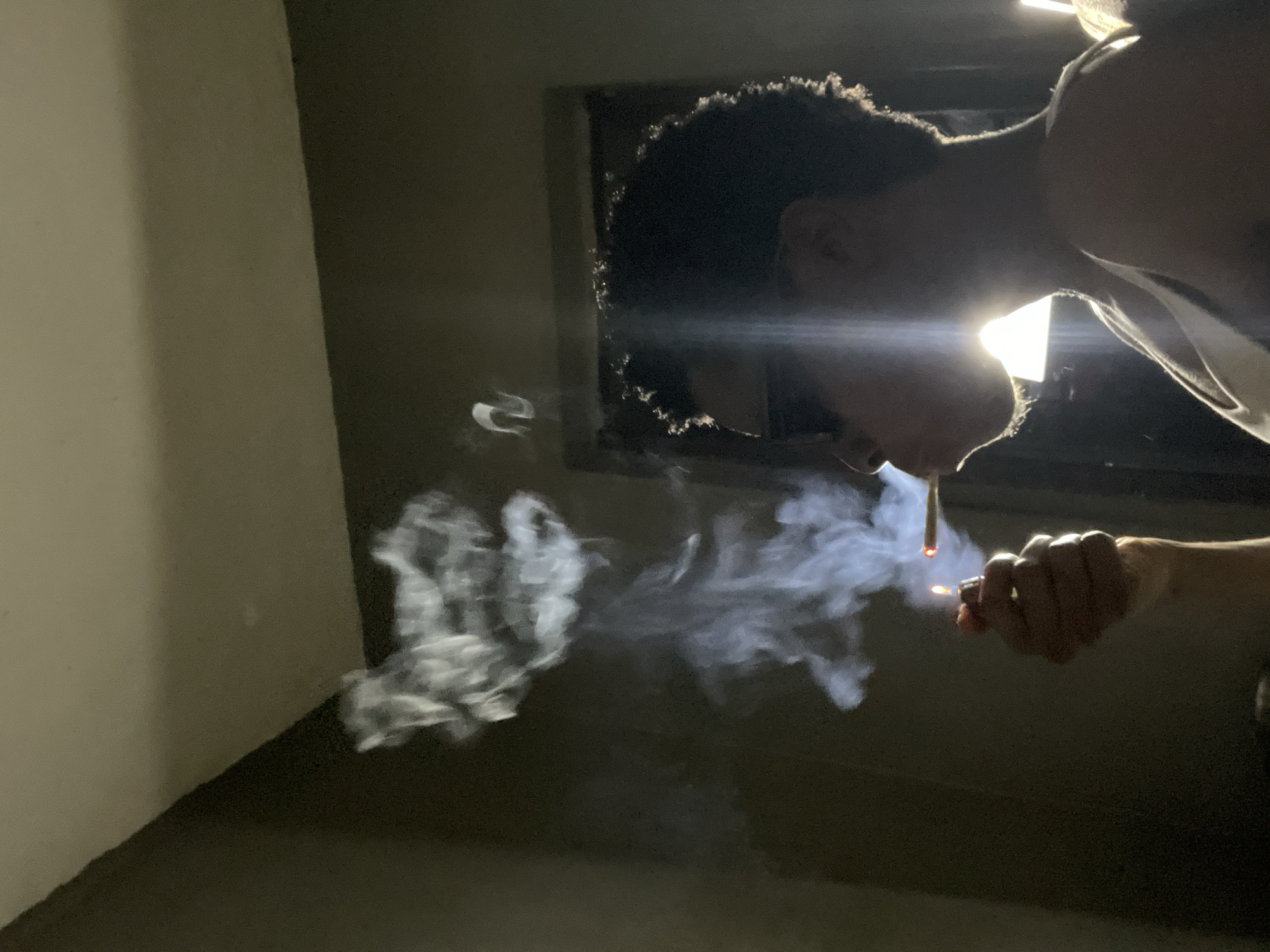 The image depicts a close-up, profile view of a person lighting a cigarette indoors, possibly in a residential setting. The lighting is dim, with a strong backlight creating a silhouette effect of the person's head and shoulders. The person's left hand is visible, holding a lighter with a flame that illuminates the cigarette. Smoke billows upwards from the cigarette and the lighter, forming abstract shapes in the air. The background is dark and indistinct, with what appears to be a doorway or window frame visible behind the person's head. The overall mood is intimate and moody, with the strong contrast between light and shadow emphasizing the act of smoking. There are no discernible elements in the image that would specifically place it in Nkpor, Nigeria.