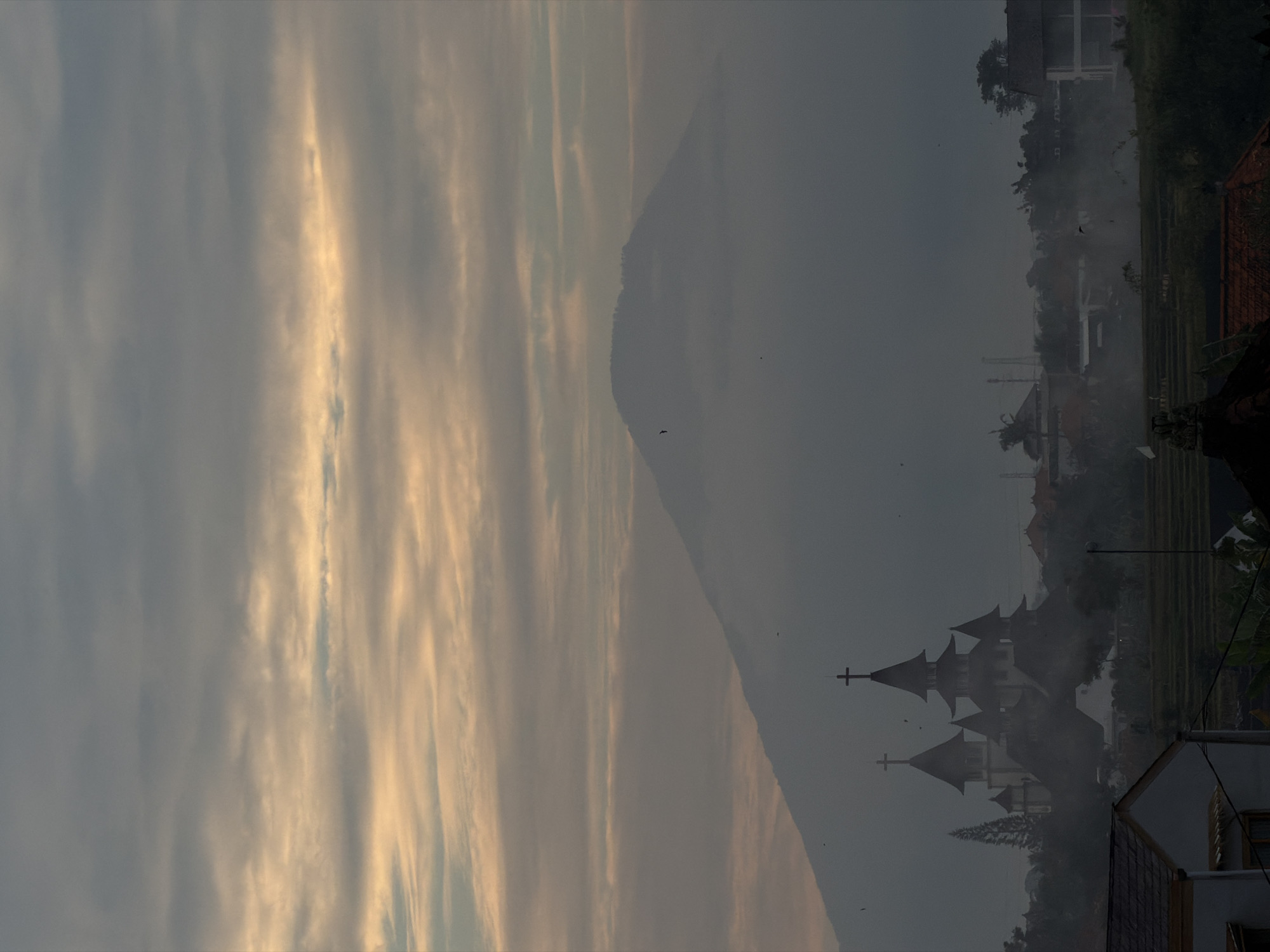 The image depicts a hazy landscape at dawn or dusk, likely in North Kuta, Indonesia. In the foreground, the roof of a house with terracotta tiles is visible on the left, and another house with a dark roof and white walls is partially seen on the right. Between these structures, a field of rice paddies is discernible. Dominating the middle ground is a cluster of buildings with distinctive, multi-tiered roofs and crosses, resembling a church or temple complex. These structures are shrouded in mist, as is the surrounding vegetation and lower slopes of a large mountain in the background. The mountain itself is partially obscured by clouds and haze, with its summit faintly visible. The sky is filled with a mix of grey and pale orange clouds, suggesting either sunrise or sunset. Small dark specks, likely birds, are scattered throughout the sky and near the mountain. The overall atmosphere is muted and serene, characterized by low visibility due to atmospheric conditions.