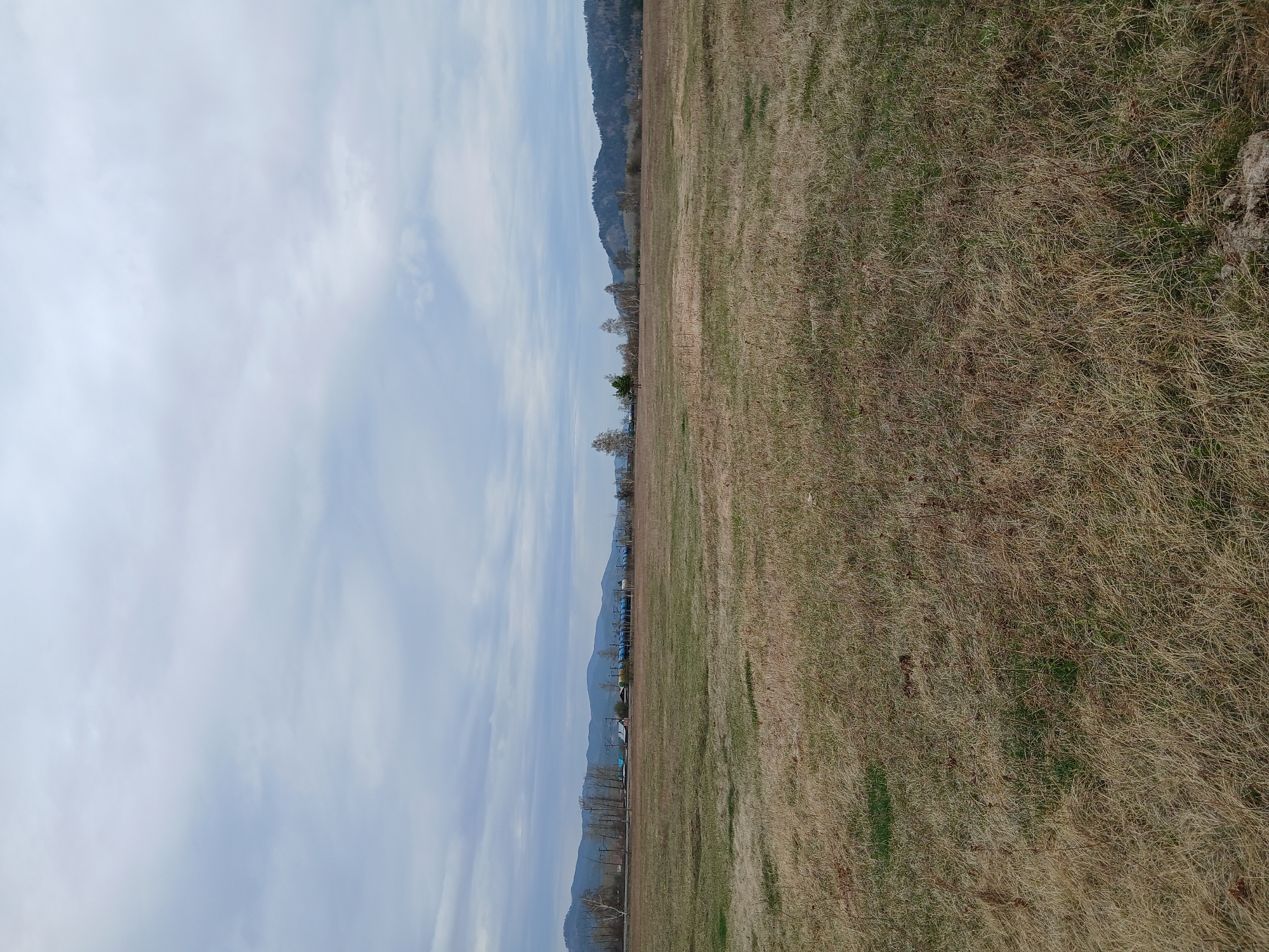 The image shows a landscape divided into two main sections: the sky and a field. The sky is light blue with scattered white clouds. The field appears to be covered with dry, brown grass. In the distance, a line of trees and hills can be seen, suggesting a mountainous or hilly backdrop. There is no information about the location as Unknown, Unknown.