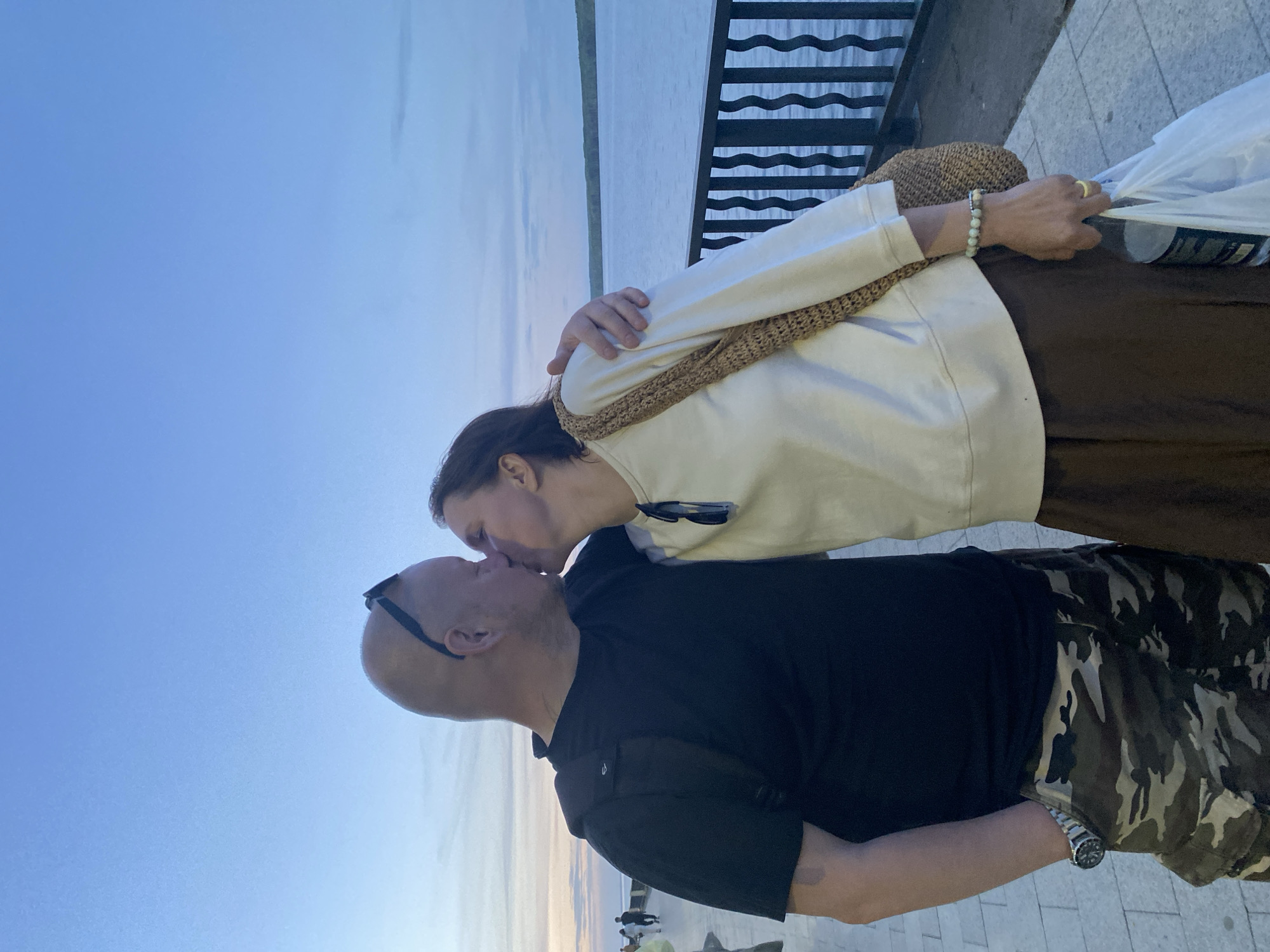 A man and a woman are kissing on a pier in Chaikovsky, Russia. The man has a bald head and is wearing a black shirt and camouflage pants. The woman is wearing a white sweater and brown skirt, and she has a straw hat hanging on her arm. They are standing next to a metal railing, and there is water in the background. The sky is blue with some clouds.