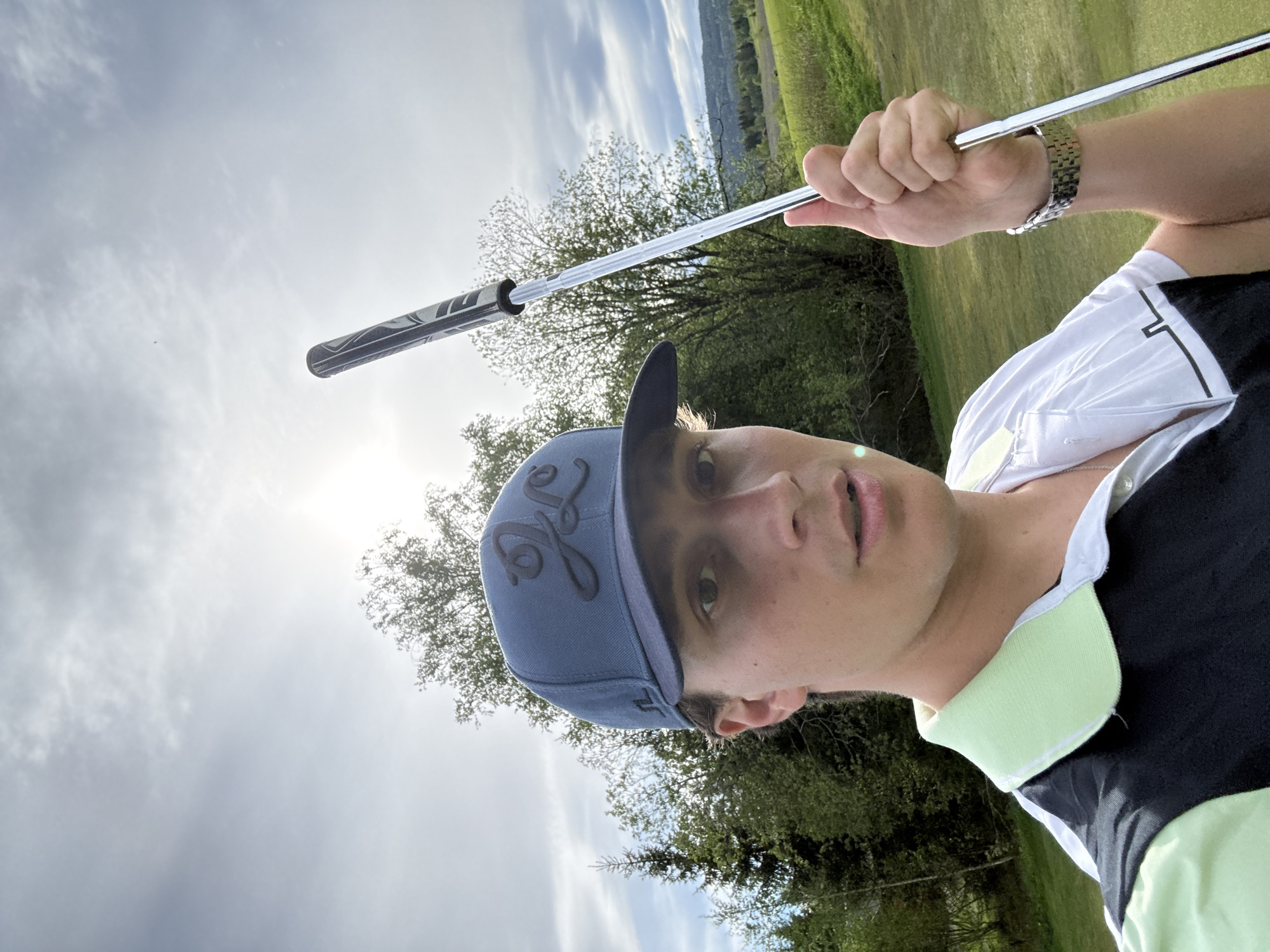 A man holds a golf club overhead in a vertical, low-angle selfie taken outdoors, likely on a golf course. He wears a blue baseball cap with a dark emblem, a white shirt with a light green collar, and a metal watch on his left wrist. Trees and a grassy field are visible in the background, along with a bright sky. The location may be Hakadal, Norway.