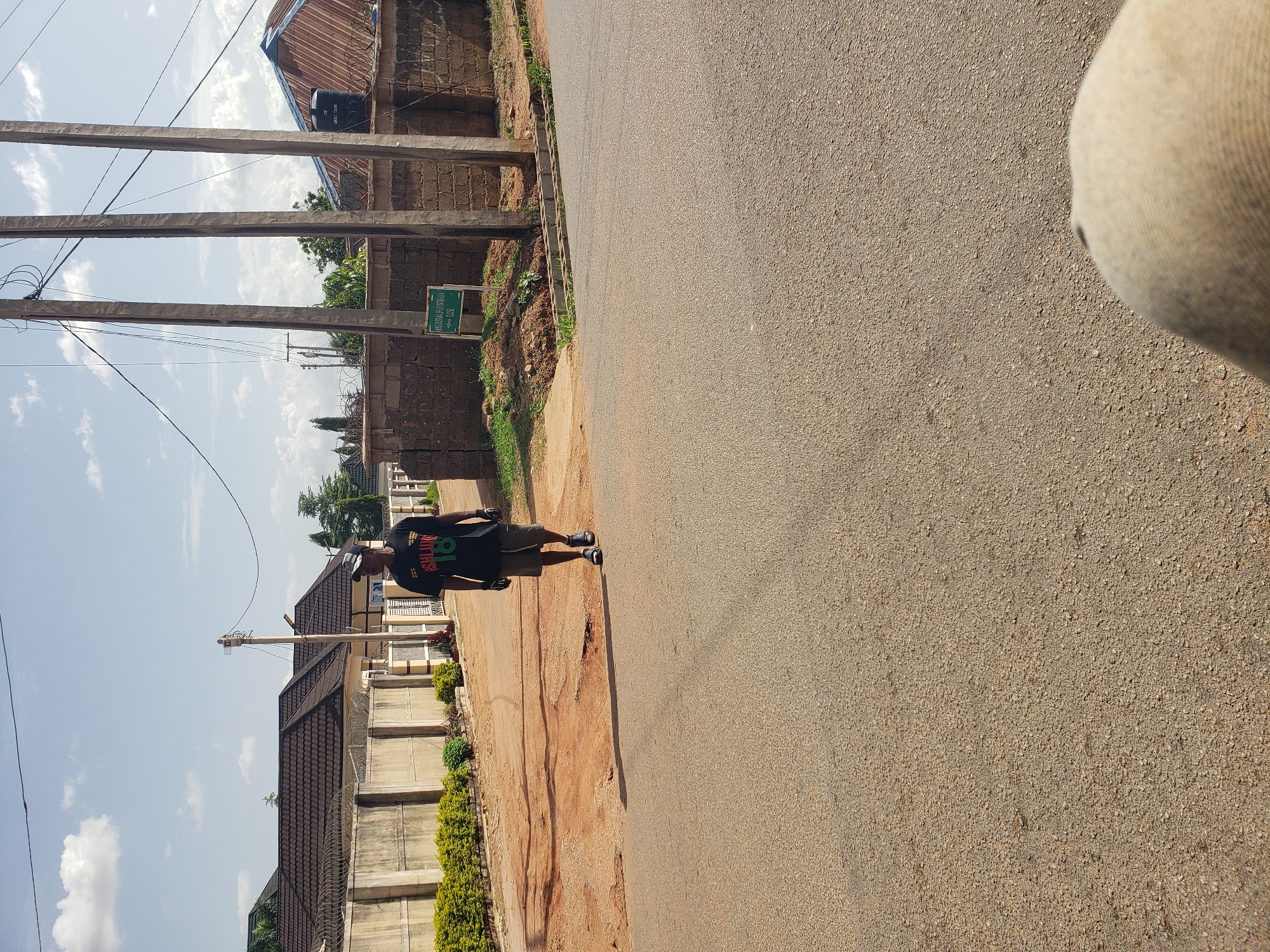 In Gura Top, Nigeria, a man walks on an asphalt road. He is wearing a black t-shirt with green and red lettering and graphics, khaki shorts, and dark slip-on shoes. He is also wearing a baseball cap. The road is bordered by a dirt path on the left and a brick wall on the right, with a green sign pointing left. Beyond the wall, there are buildings with corrugated metal roofs and barbed wire fencing. The sky is partly cloudy, and there are several utility poles with wires running between them. The scene appears to be in a residential area.