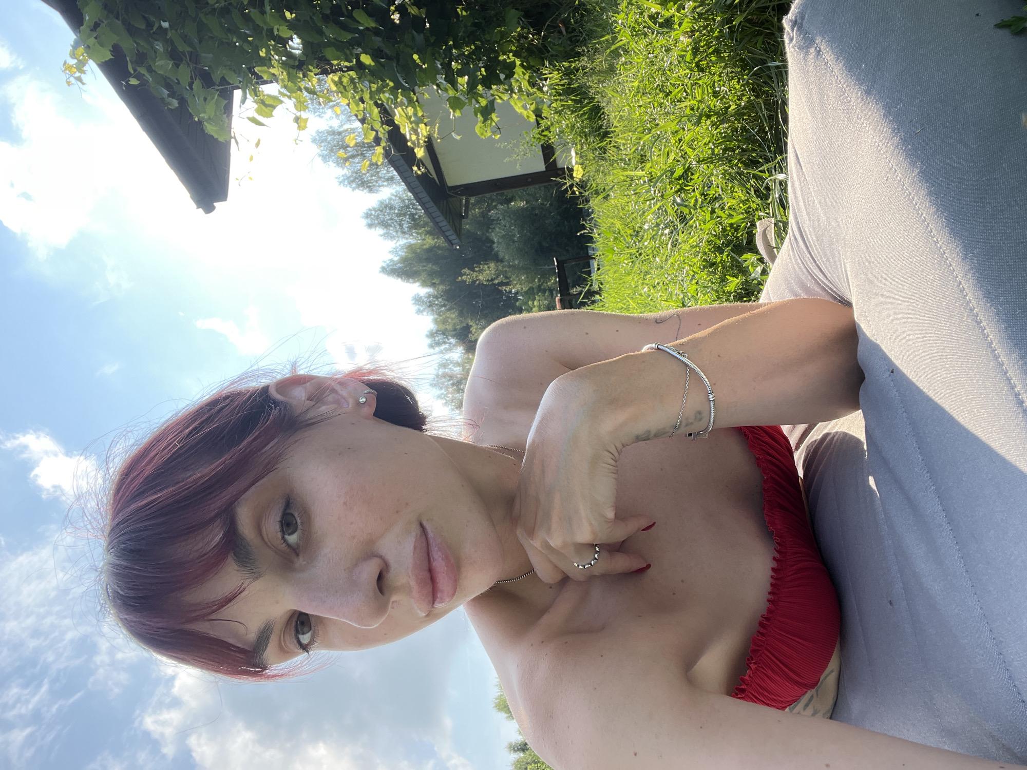 In Нагорный, Russia, a young woman with auburn hair, wearing a red bikini top and light-colored pants, is captured in a close-up shot. She is lying down, with her head resting on her arm, and her hand is touching her chin. The background features a lush green field, trees, and a house. The sky is bright blue with white clouds, and the sun is shining, creating a warm and inviting atmosphere. The woman appears to be relaxed and enjoying the outdoors.