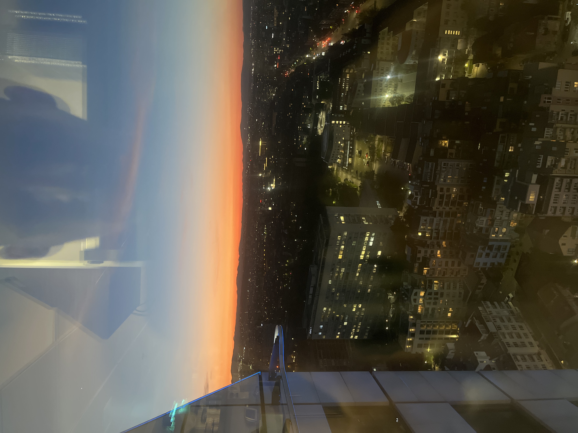 The image shows a nighttime view of the city of Sofia, Bulgaria, from a high vantage point, possibly a tall building or skyscraper. The sky transitions from a dark blue to orange, suggesting either sunset or sunrise. Below, the city is illuminated by streetlights and lights from buildings. Numerous buildings of varying sizes and architectural styles are visible, some with windows lit. Streetlights indicate the presence of roadways and traffic, although the traffic itself is difficult to discern. There is the outline of a mountain range on the horizon. The picture appears to be taken from behind a glass barrier/ledge, suggesting it is an interior shot.