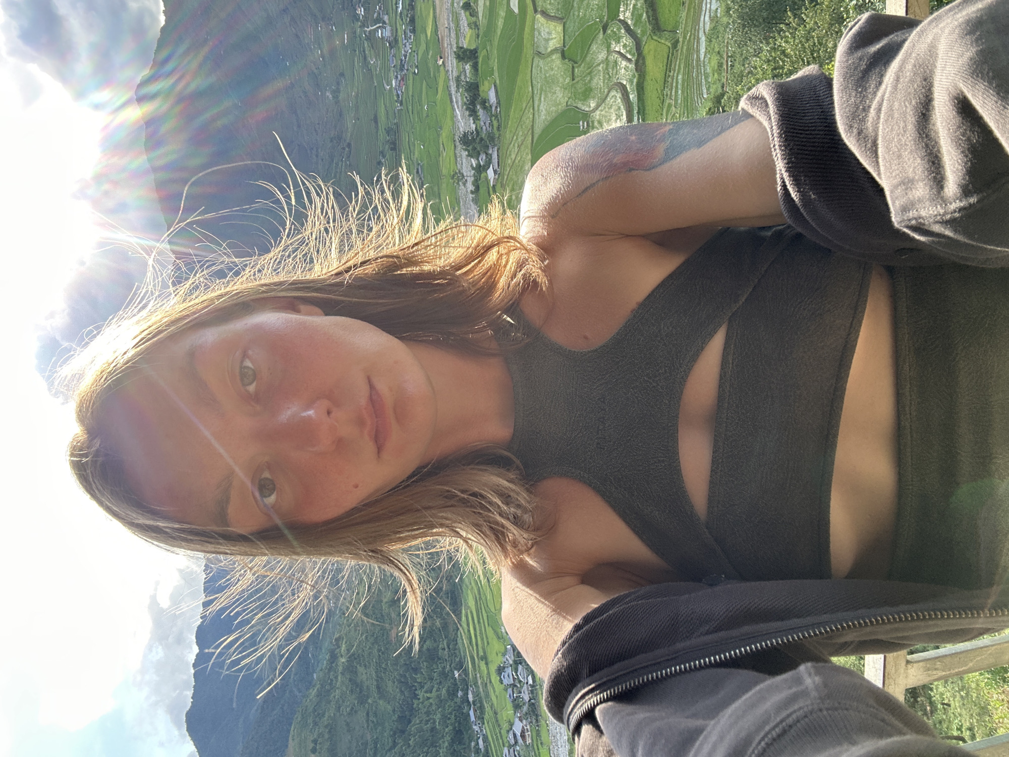 A female subject, appearing to be in her 20s or 30s, takes a selfie outdoors in Tú Lệ, Vietnam. She has medium-length light brown hair, appearing wind-blown and backlit, creating a bright halo and distinct strands around her head. Her face shows some redness, and a small mole is visible below her left eye. She wears a dark, possibly olive green, halter-neck top with a cut-out design across the chest and midriff, paired with matching dark bottoms. A dark grey zip-up hoodie is worn unzipped, with its sleeves pulled up on her arms. A colorful tattoo is visible on her right upper arm. The subject gazes directly into the camera.
The background reveals a expansive mountainous landscape dominated by vibrant green terraced rice paddies that curve along the valley floor and mountain slopes. A small village with multiple residential structures is nestled within the valley, adjacent to what appears to be a river or stream. The scene is brightly lit by strong sunlight from behind the subject, causing lens flare and distinct rainbow-colored sun rays to appear, particularly on the right side of the image. The sky is partly cloudy with patches of blue. A metal railing is partially visible in the bottom left foreground, suggesting the subject is positioned on an elevated viewpoint overlooking the valley.