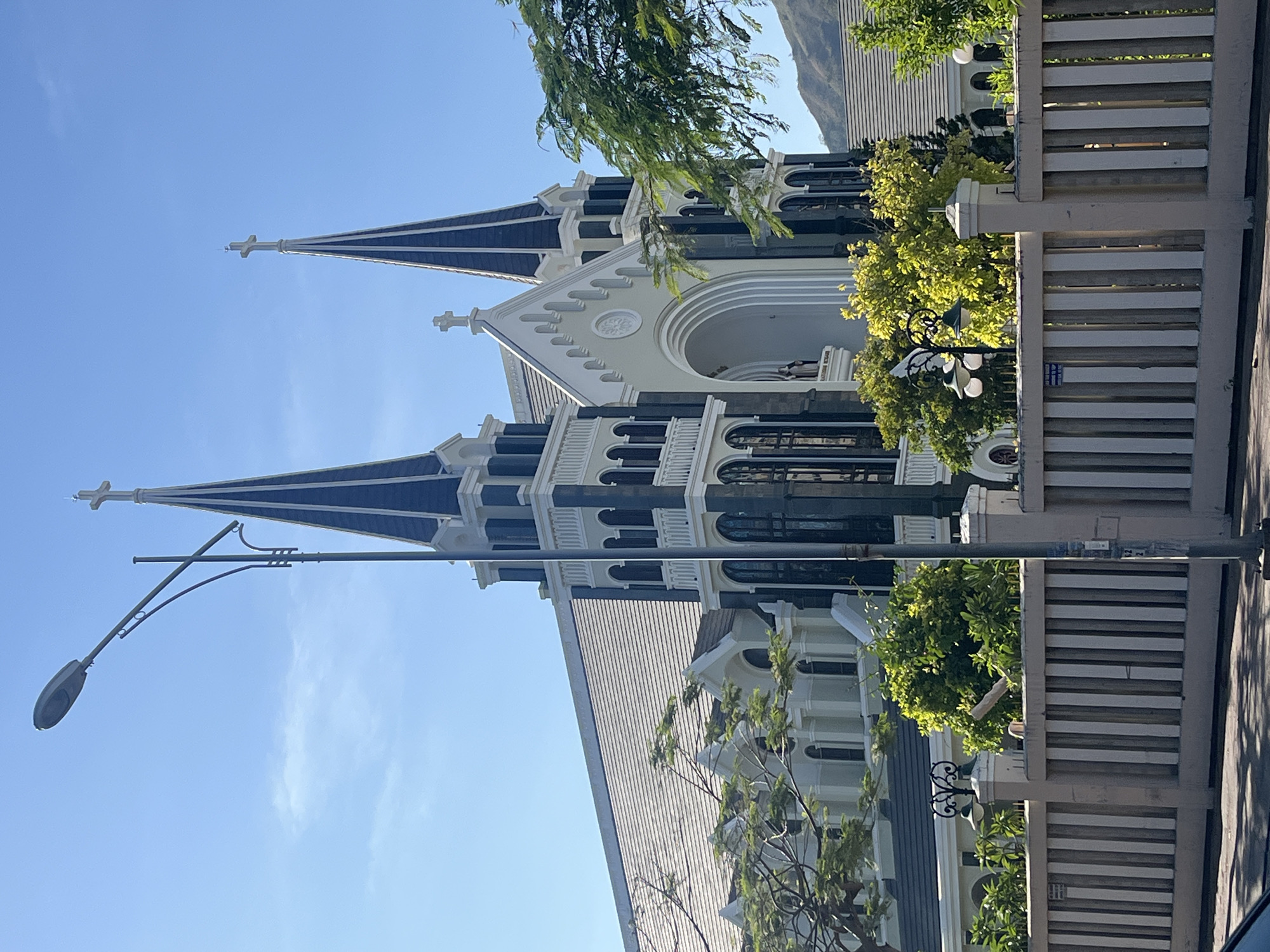 The image depicts a church in Bac Nha Trang Ward, Vietnam. The church features two prominent spires with crosses atop, a gray roof, and arched windows. A street lamp is visible in the foreground, along with a fence partially obstructing the view. The sky is clear and blue. Greenery is present in the form of trees and shrubs. A mountain is visible in the background.
