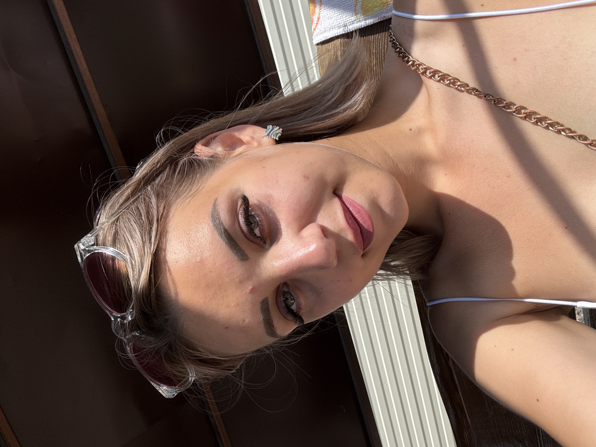 A young Caucasian woman with blonde hair is pictured in a close-up, outdoor shot. She is wearing a white tank top and a gold chain purse strap is visible across her shoulder. She has on pink lipstick and is wearing a silver earring. Sunglasses are resting on top of her head. The background consists of horizontal white blinds and wooden slats, suggesting an outdoor seating area or shelter. The lighting indicates it is daytime, with sunlight casting shadows on her skin. The location is a public park in Krasnogorskoye, Russia.