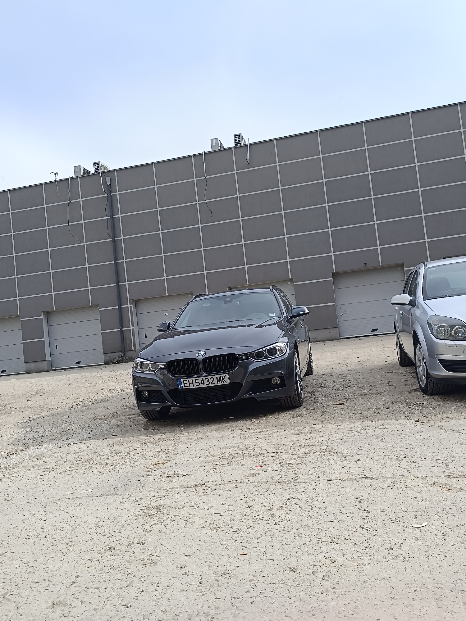 The image presents an outdoor scene in what appears to be an industrial or commercial area. Two cars are visible: a dark gray BMW wagon with license plate EH5432MK and a silver Opel Astra. They are parked on a light-colored gravel or dirt surface. In the background is a large, gray building with a grid-like pattern and multiple garage doors. The sky is overcast. This is likely a suburb of Pleven, Bulgaria.