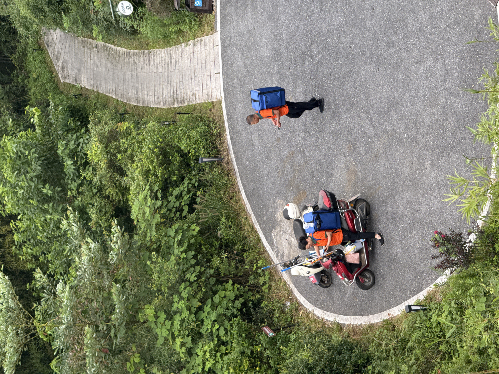 In Wuxing District, China, two individuals are observed in a paved area surrounded by dense green foliage. To the left, a person wearing an orange shirt and black pants stands next to a red scooter. The scooter has a large blue insulated bag mounted on the rear, and there are other items secured to the scooter, including a white helmet and what appears to be a bucket. The person is holding a mobile phone and seems to be interacting with it. Behind this scooter is another scooter, primarily white and red, also equipped with a blue insulated bag. Further to the right, a second person, also in an orange shirt and black pants, walks away from the scooters, carrying a large blue insulated bag on their back and holding a mobile phone in their hand. A paved pathway with stone steps winds through the greenery in the background. Signs are visible on the left and right sides of the pathway. A waste bin is located near the right side of the pathway. The overall scene depicts a delivery or courier activity in a semi-urban, green environment.
