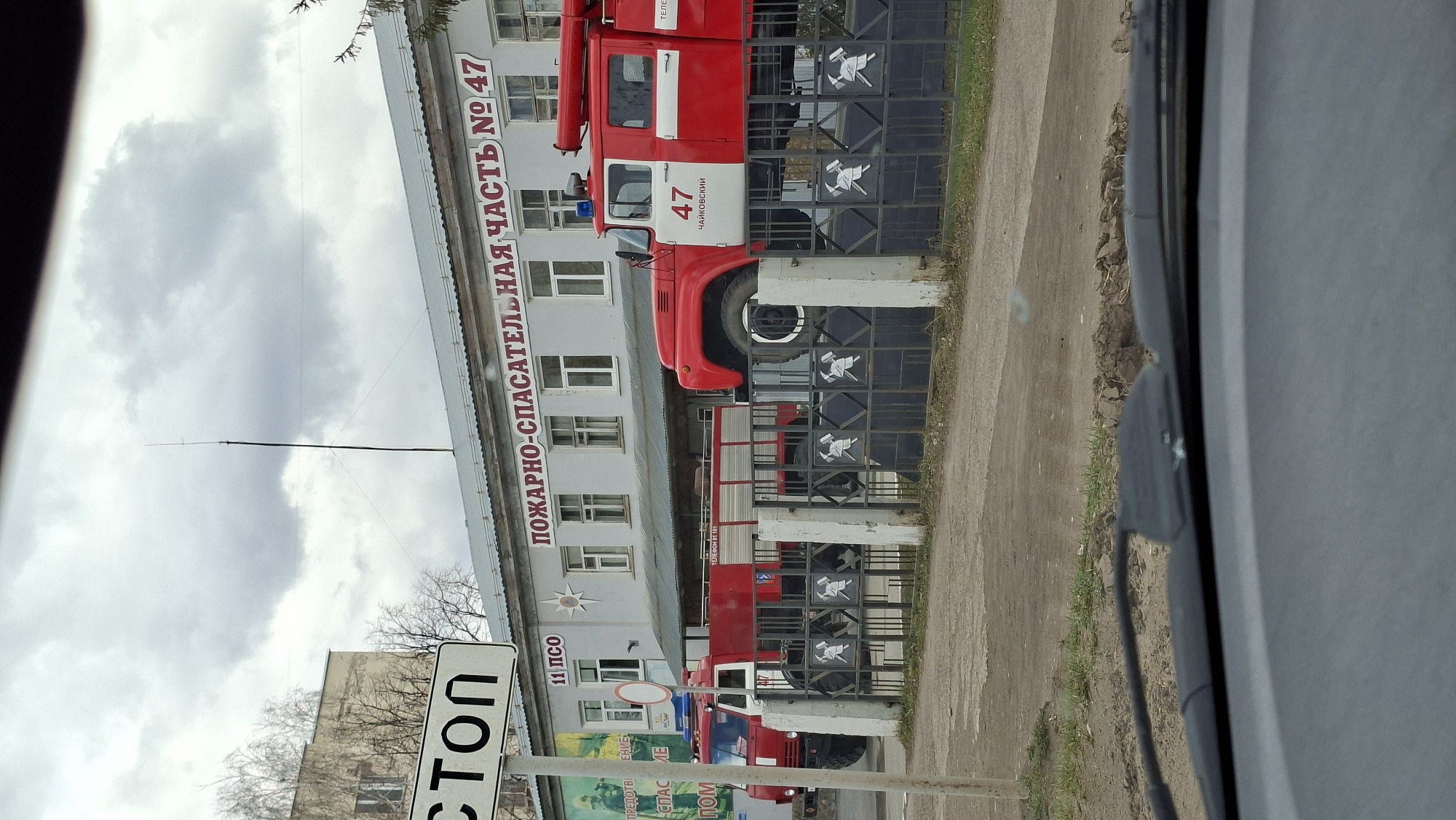 This media file captures a static view of a fire and rescue station in Chaikovsky, Russia, on an overcast day. The primary subject is a two-story, light-colored building housing the fire department, with at least two red and white fire trucks parked in front, visible through a metal fence.
The building features several windows and a large red sign across its facade. The text on this sign clearly reads "ПОЖАРНО-СПАСАТЕЛЬНАЯ ЧАСТЬ № 47" (Fire and Rescue Unit No. 47). Below this, a smaller sign with "11 ПСО" (likely indicating a fire-rescue squad) is visible. Another sign on the building displays "ТЕЛЕФОН 01 101," providing emergency contact numbers.
One prominent fire truck, red with a white stripe, has the number "47" and the word "ЧАЙКОВСКИЙ" (Chaikovsky) on its side, confirming the location. The vehicle is equipped with a blue light on its roof. A second fire truck is partially visible further down.
A dark metal fence with decorative elements, including stylized axes and helmets, separates the fire station's property from a paved area or road. Beyond the fence, there is a dirt and grass shoulder. To the left, a "СТОП" (STOP) sign is visible. The sky is largely covered with thick, grey clouds, suggesting an overcast day. Bare tree branches can be seen in the background, implying a non-summer season. The image appears to be taken from a vehicle, with part of the car's interior visible on the right side. A billboard with partial Russian text like "ПРЕДОТВРАЩЕНИЕ" (Prevention), "СПАСЕНИЕ" (Rescue), and "ПОМОЩЬ" (Help) is also visible near the STOP sign.