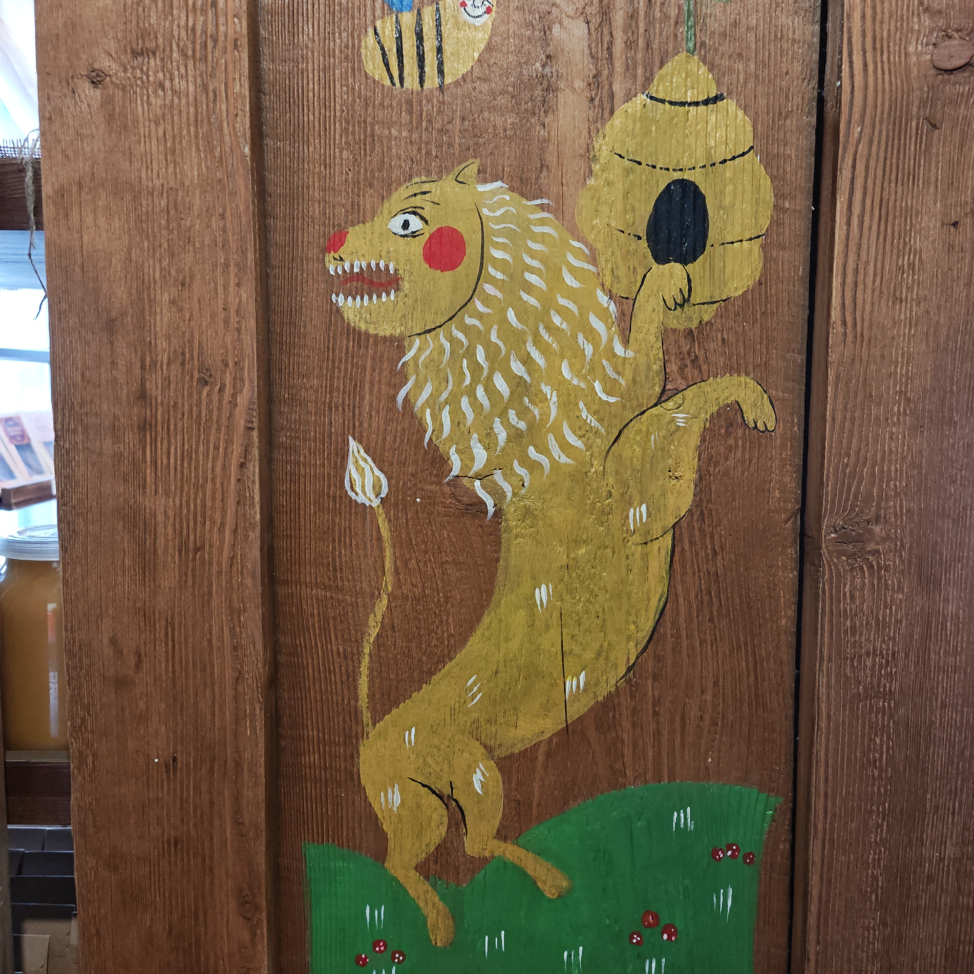A wooden panel displays folk art featuring a yellow lion holding a beehive, standing on green grass with red mushrooms. Above the lion is a smiling bee. The panel appears to be part of a larger structure, likely a door or wall. The image appears to be taken indoors, possibly in Ново-Никольское, Russia, though this location information is based on the prompt and not visible in the image itself.