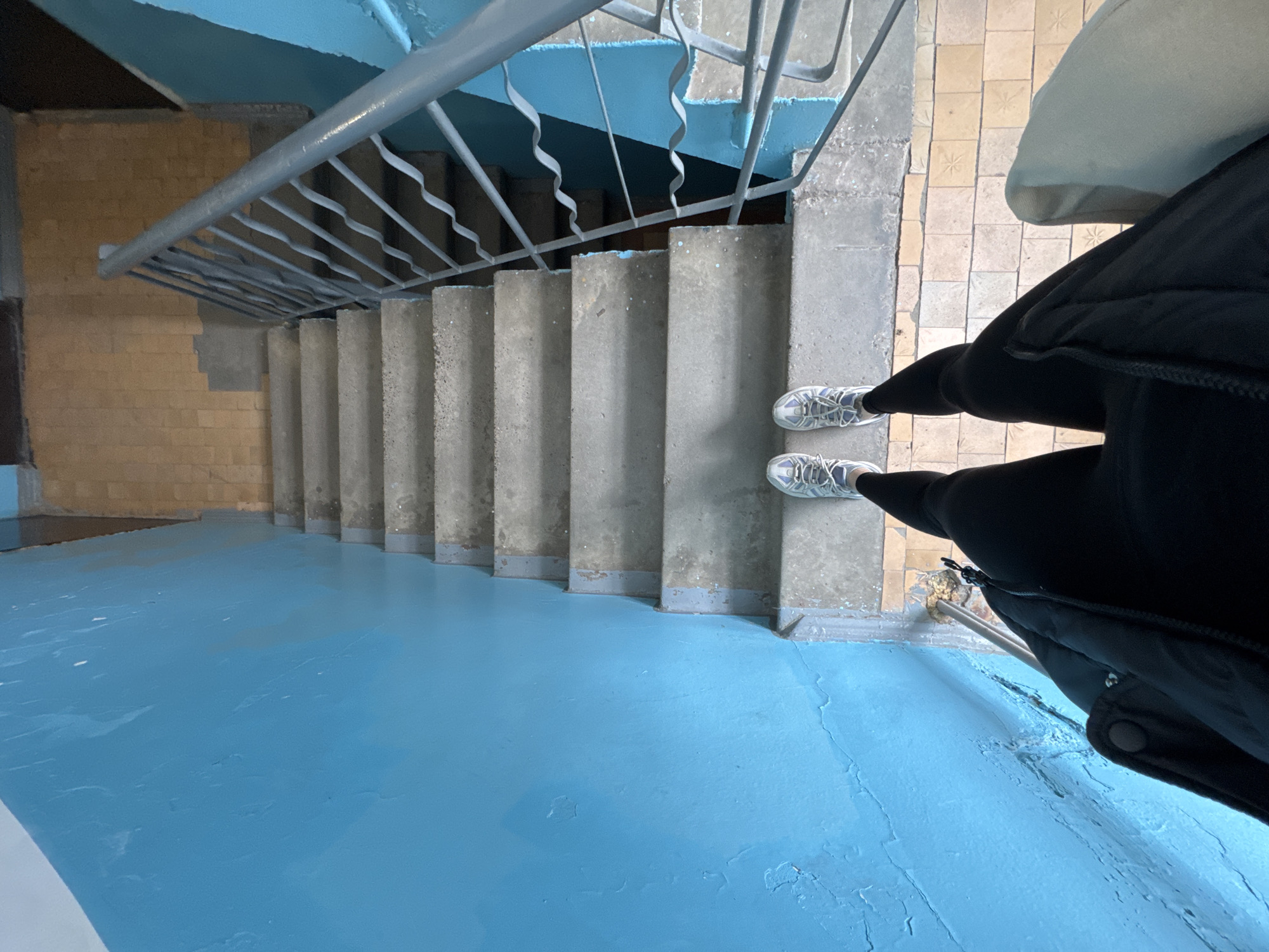 View looking down a stairwell painted light blue. The stairs themselves are concrete and the handrail is grey. An individual in black leggings and white sneakers stands on the top step, wearing a black puffer jacket with a hood. The ceiling is light blue. A wall to the left is yellow brick. Some tile flooring is visible on the right, near the top of the stairs. No evidence supports the location of Chaikovsky, Russia.