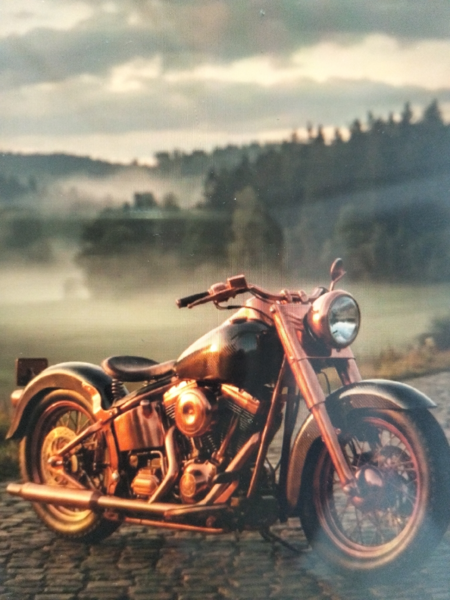 The image depicts a vintage copper-colored motorcycle parked on a cobblestone surface. The motorcycle is positioned in the foreground, angled slightly towards the viewer's left. The background features a misty, forested landscape under a cloudy sky. The scene has a soft, atmospheric quality, with fog obscuring the details of the distant trees and hills. There are no people or discernible actions in the image. The location is specified as the city of Payagaboh, Indonesia, though the image itself does not contain any visual cues to confirm this specific geographical context.