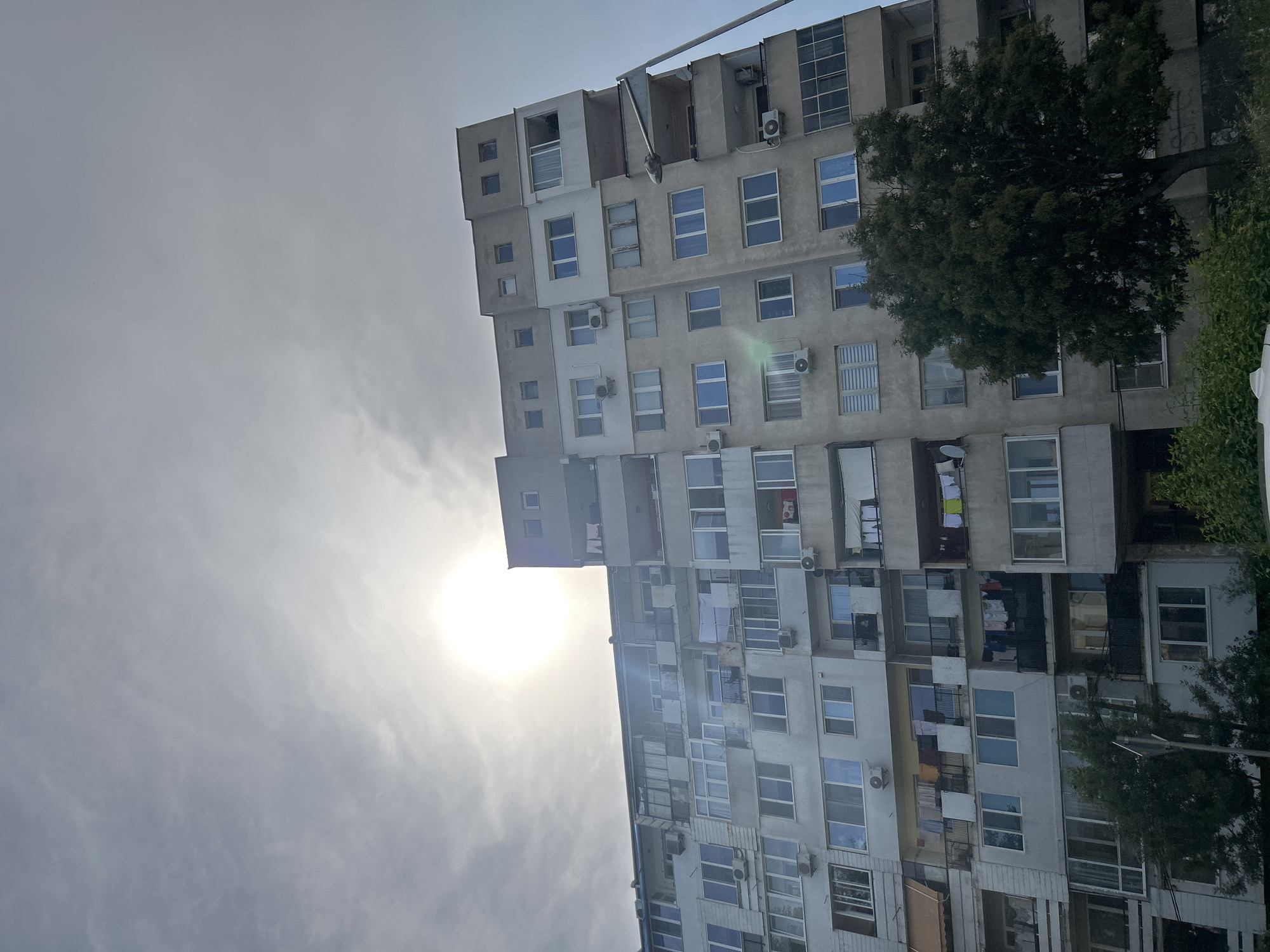 The image depicts a multi-story residential apartment building in Varna, Bulgaria, viewed from a low angle. The sun is positioned brightly in the upper-center sky, directly behind and slightly above the building, causing significant glare and overexposure in that area. The sky is partially cloudy with a diffused haze around the sun.
The building is constructed primarily of light-colored concrete, exhibiting various shades of grey and beige across its facade. It features numerous windows and balconies. Several balconies are enclosed with glass or other materials, while others remain open. External air conditioning units are mounted on the exterior walls of multiple apartments. Clothes are visible drying on lines on several open balconies across different levels, indicating residential occupancy. A small satellite dish is affixed to one balcony. The building's architecture suggests a typical post-socialist residential block.
In the foreground, a large, dark green deciduous tree with dense foliage partially obscures the lower-left and central sections of the building. To the right, a grey street light pole extends upwards, with its luminaire positioned roughly at the fifth-story level. No individuals are visible within the frame. The immediate foreground shows some low green vegetation.